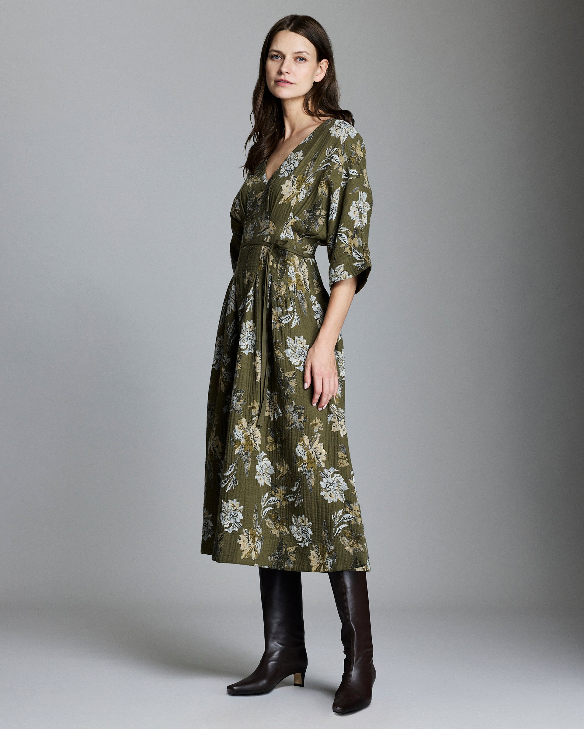 A woman wearing a long, olive green floral dress with a V-neck and three-quarter sleeves stands in profile. She pairs the dress with knee-high brown boots and has long, straight hair. The background is plain gray.