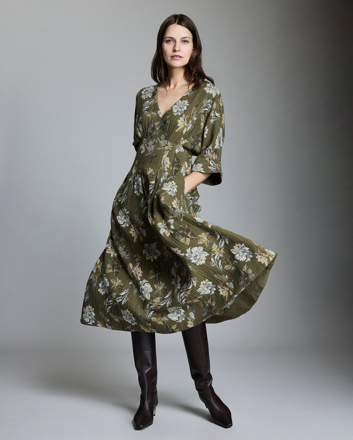 A woman stands in a studio wearing a long, olive-green floral dress with a v-neck and elbow-length sleeves, paired with tall dark brown boots. She has long brown hair and a neutral expression. The background is gray.