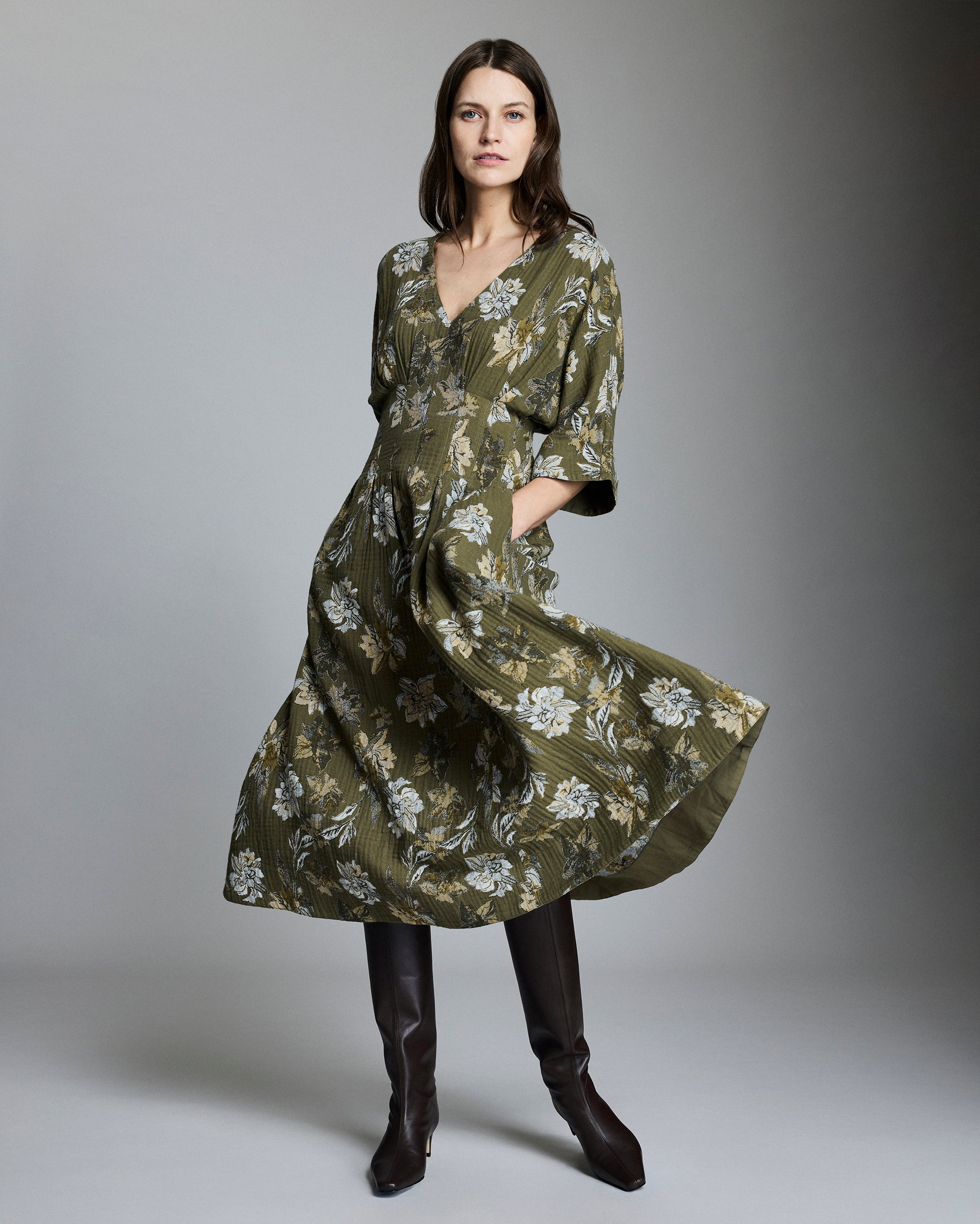 A woman stands in a studio wearing a long, olive-green floral dress with a v-neck and elbow-length sleeves, paired with tall dark brown boots. She has long brown hair and a neutral expression. The background is gray.