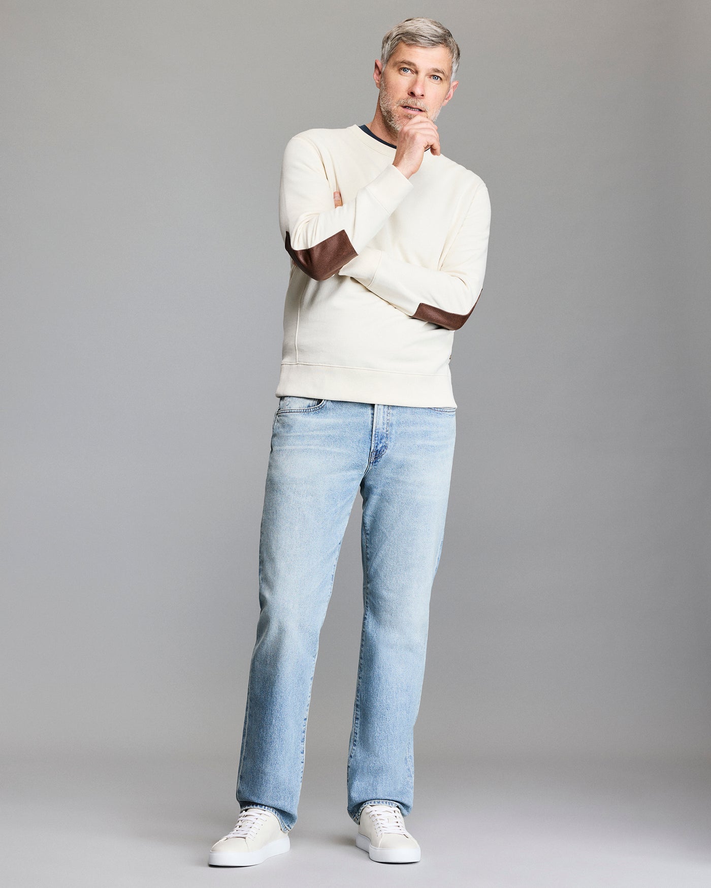 A man with gray hair and a beard stands thoughtfully against a plain background, wearing a cream sweater with brown elbow patches, light blue jeans, and white sneakers.