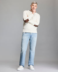A man with gray hair and a beard stands thoughtfully against a plain background, wearing a cream sweater with brown elbow patches, light blue jeans, and white sneakers.