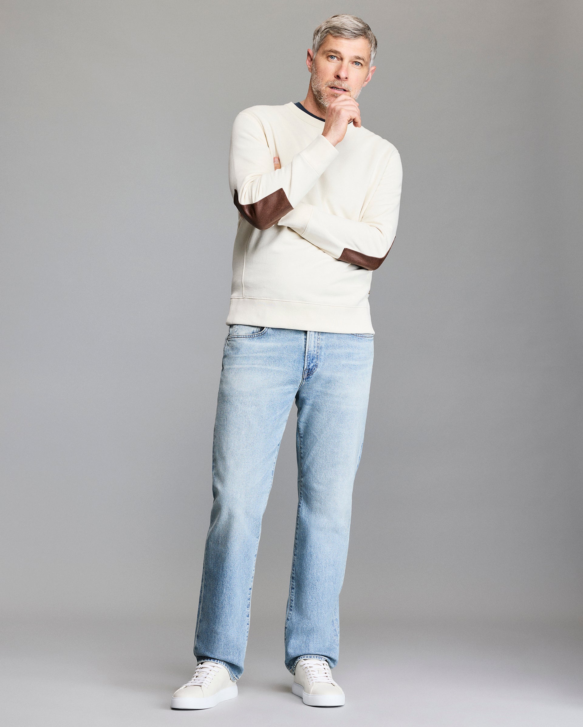 A man with gray hair and a beard stands thoughtfully against a plain background, wearing a cream sweater with brown elbow patches, light blue jeans, and white sneakers.