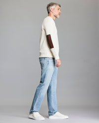 A man with gray hair stands in profile against a gray background, wearing a cream-colored sweater with a brown elbow patch, light blue jeans, and white sneakers.