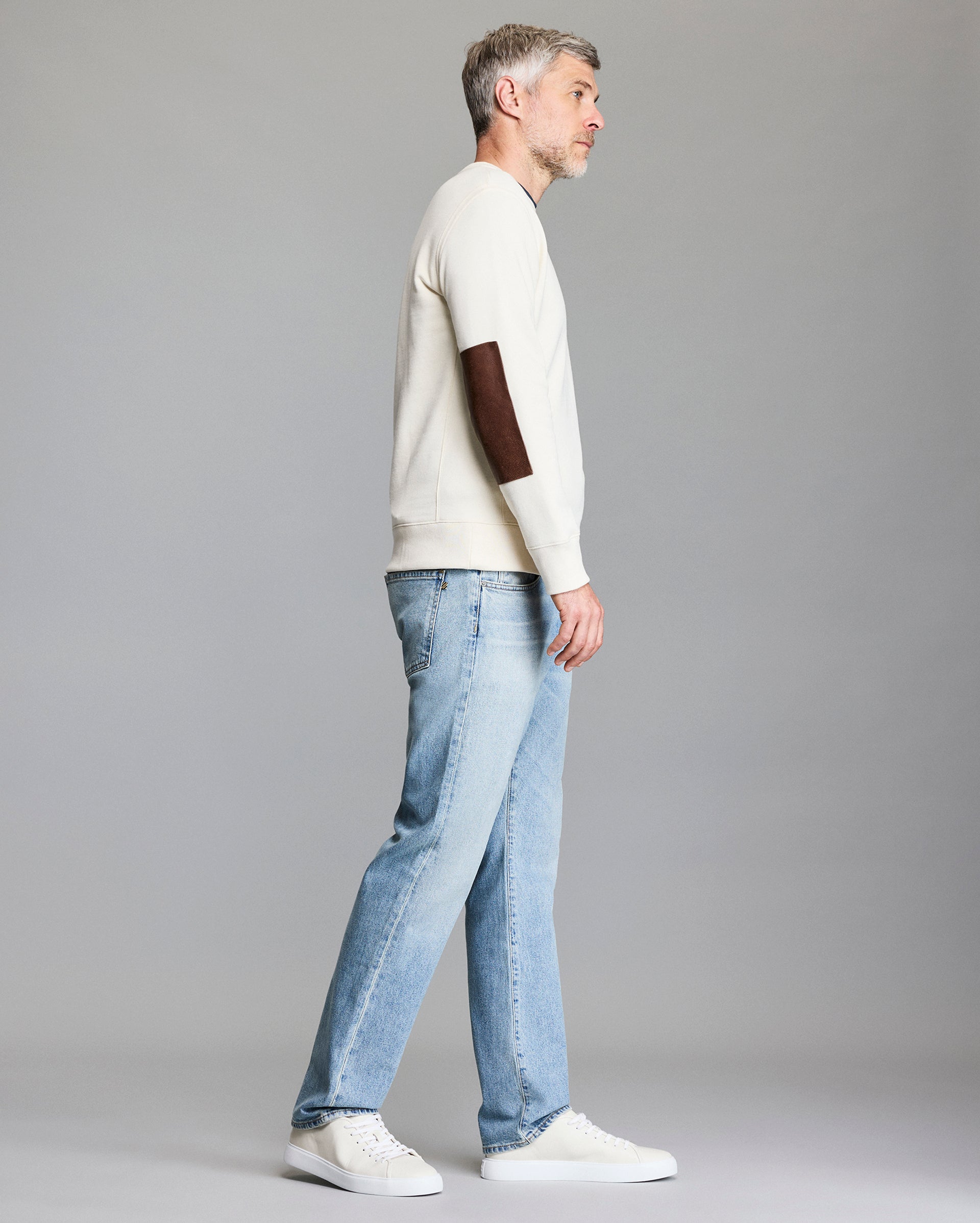 A man with gray hair stands in profile against a gray background, wearing a cream-colored sweater with a brown elbow patch, light blue jeans, and white sneakers.