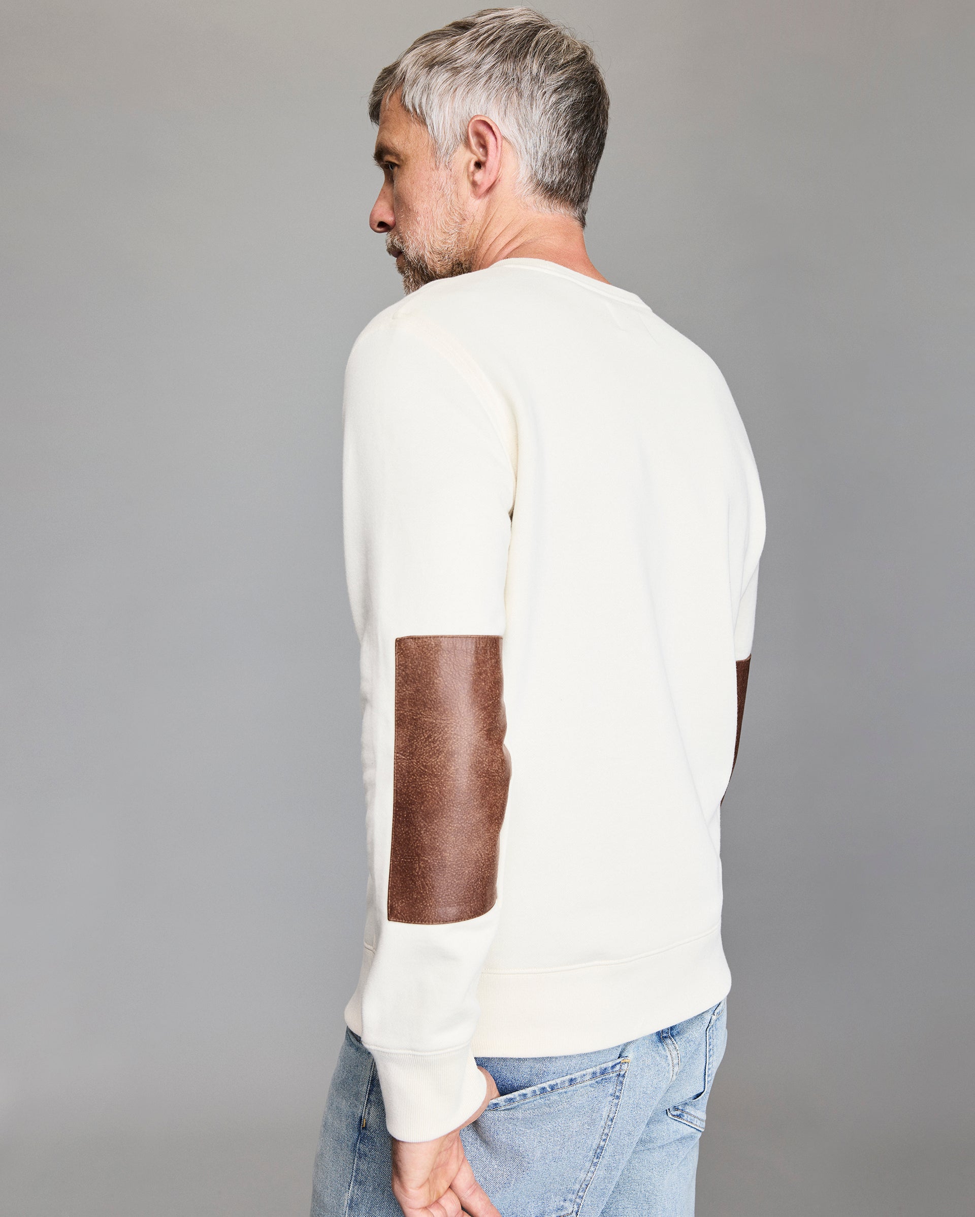 A man with short gray hair, wearing a cream-colored sweatshirt with brown suede elbow patches and light blue jeans, stands facing away from the camera against a plain gray background.