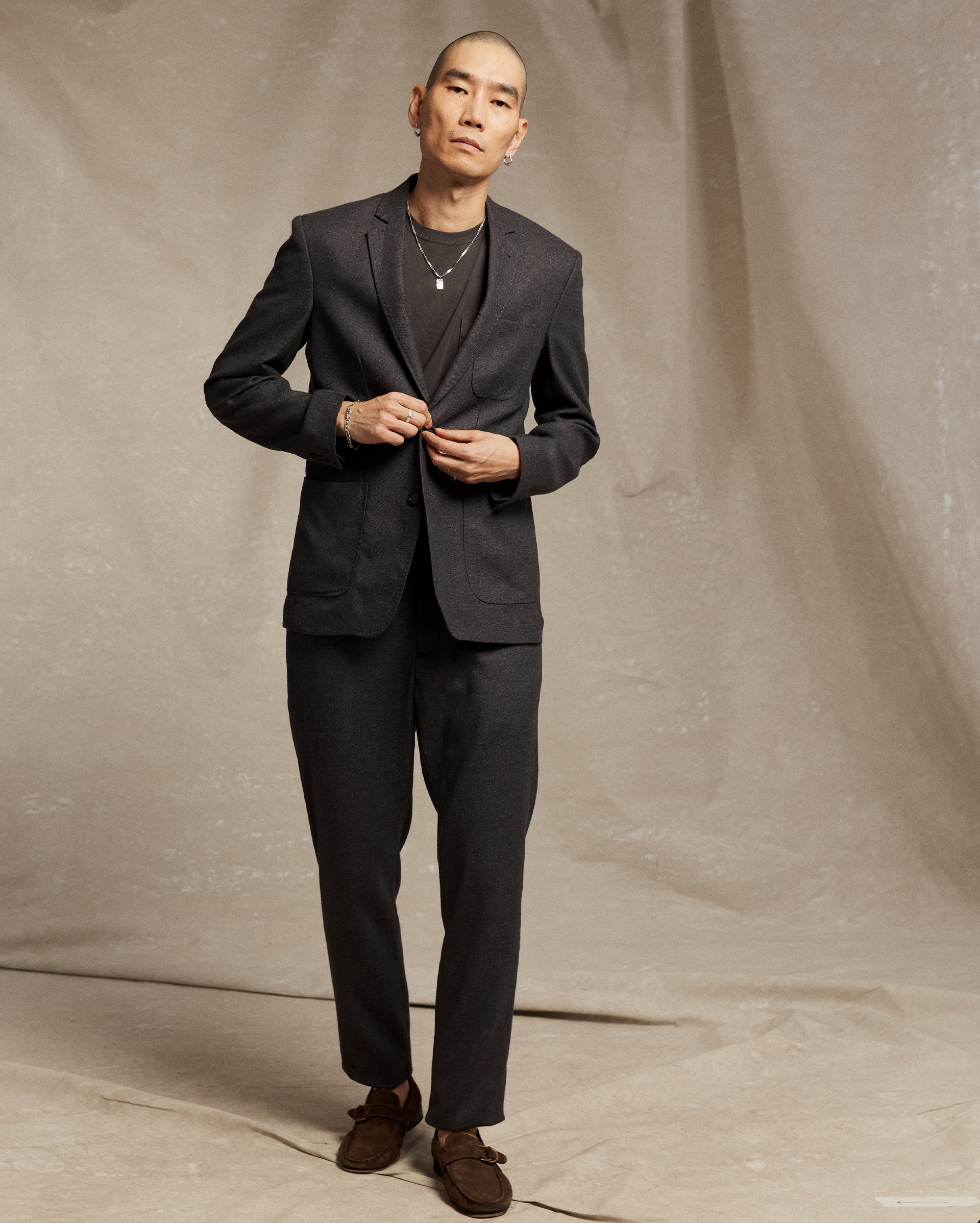 A man with a shaved head wears a dark suit over a black shirt, brown loafers, and simple jewelry. He stands against a beige, textured backdrop, buttoning his suit jacket while looking directly at the camera.