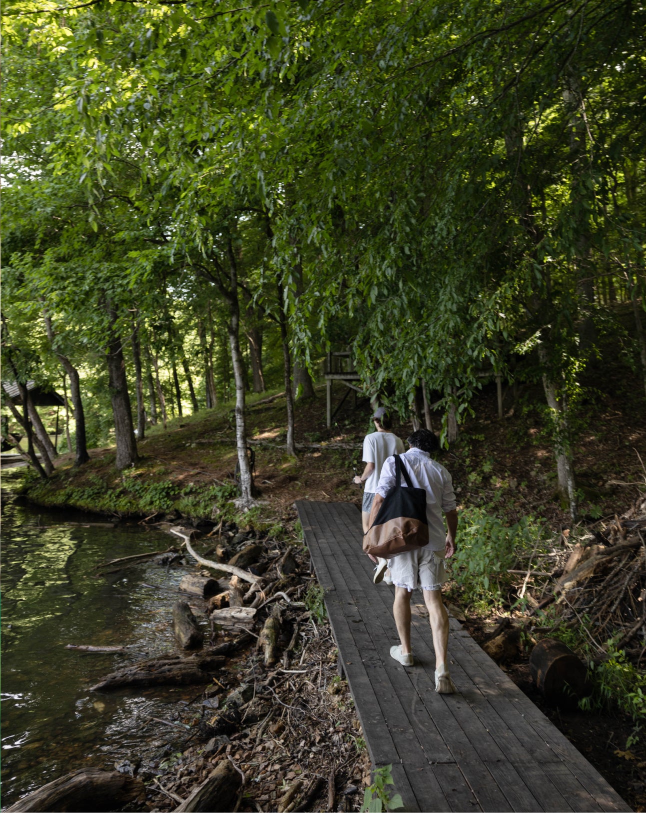 Two men walk across a wooden bridge into woods by the lakeside.
