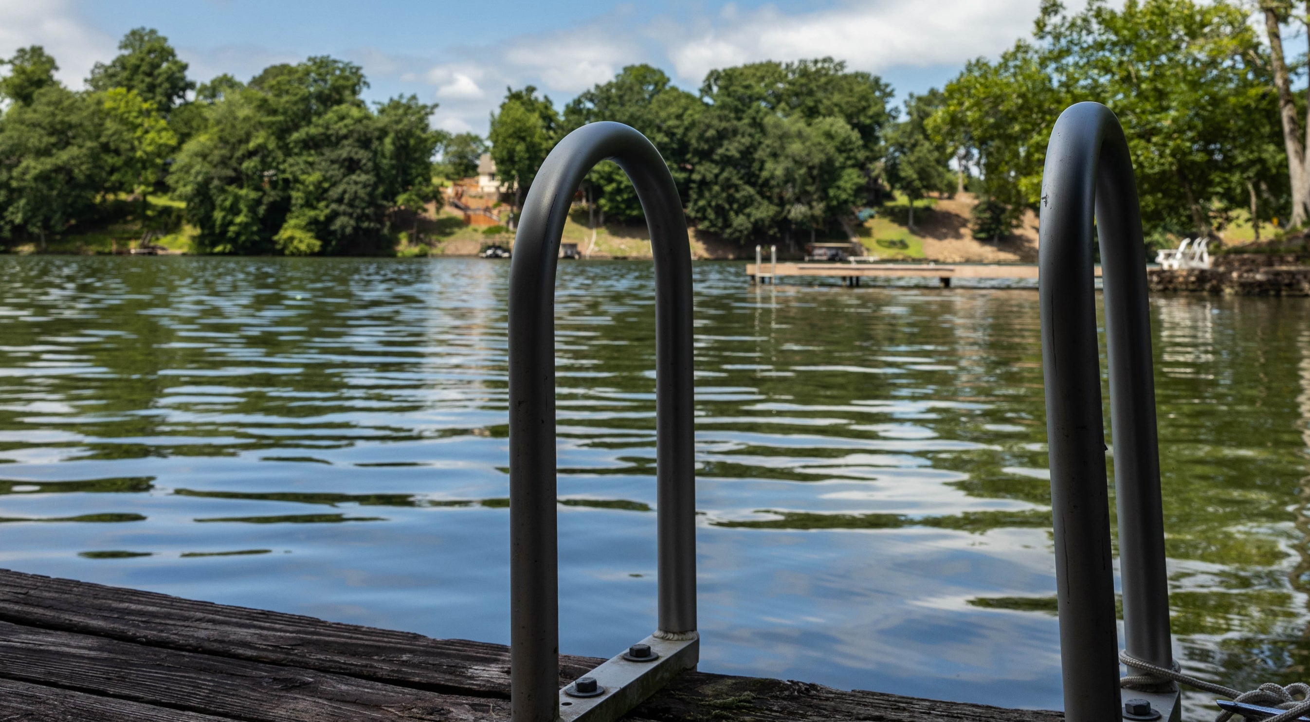 A black iron ladder mounted on a wooden deck leads into a lake with a warm summer day in the background.