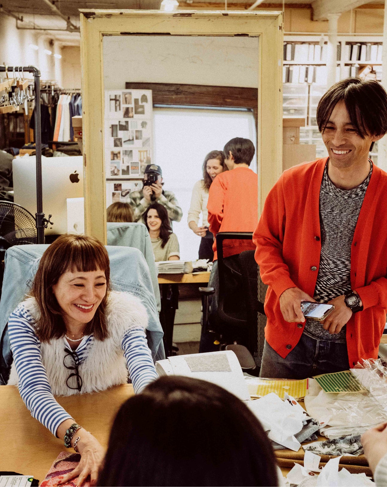 Two people smiling in a workshop or office setting with various items and people in the background.