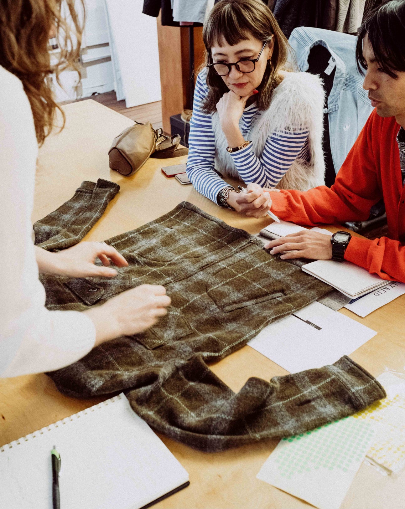 Three people examining plaid fabric on a table with notes and a phone.
