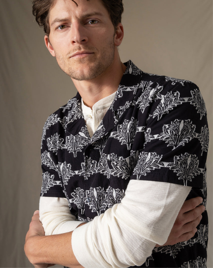Man wearing a black and white patterned shirt over a white long-sleeve shirt against a beige background