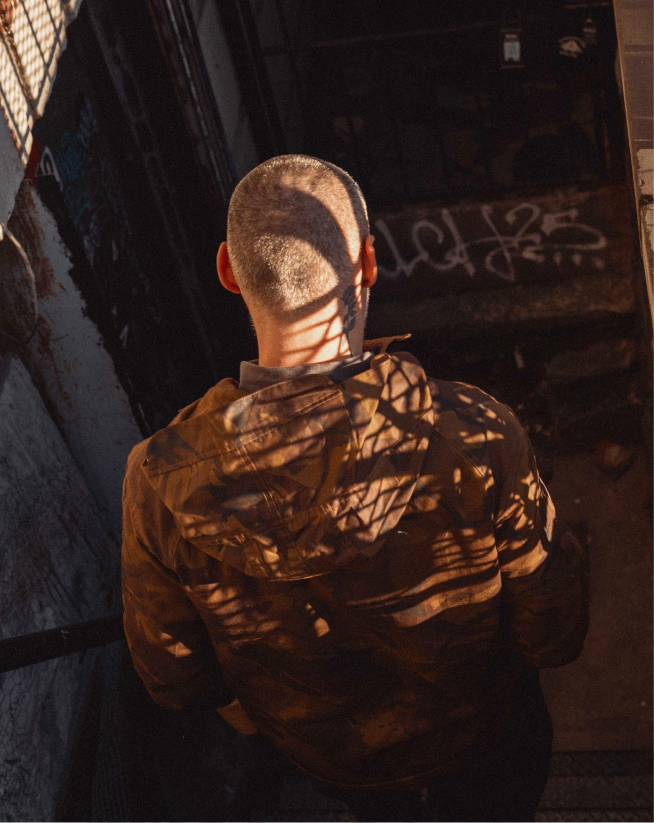 Person wearing a brown jacket with a textured pattern, standing in front of a dark wall with graffiti.