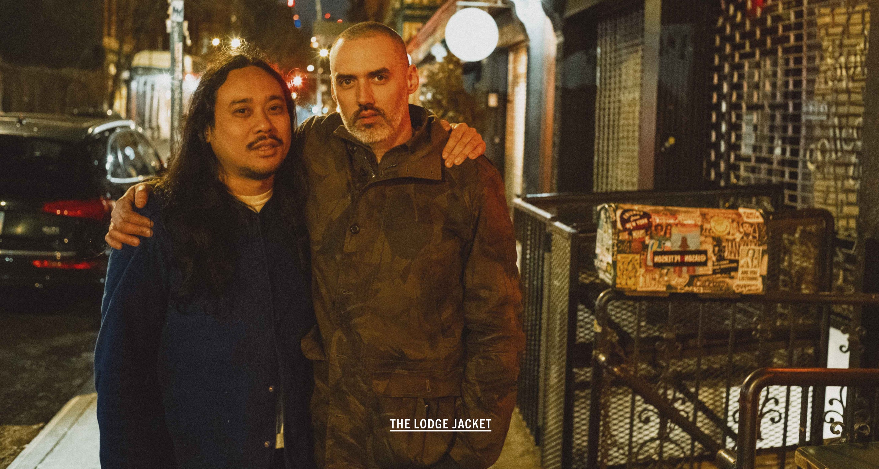 Two men standing together on a street at night with a store in the background.