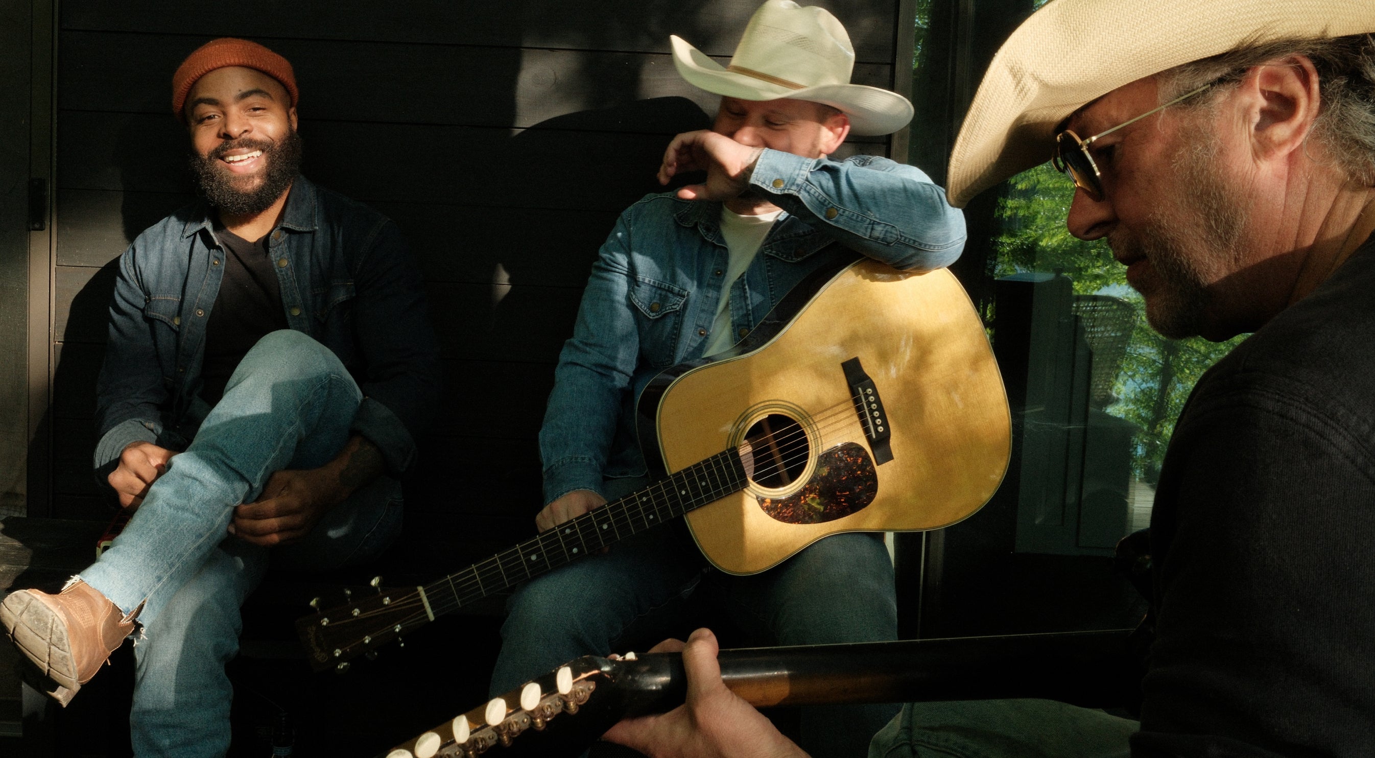 Three musicians playing guitar and harmonica in a vehicle with a scenic background
