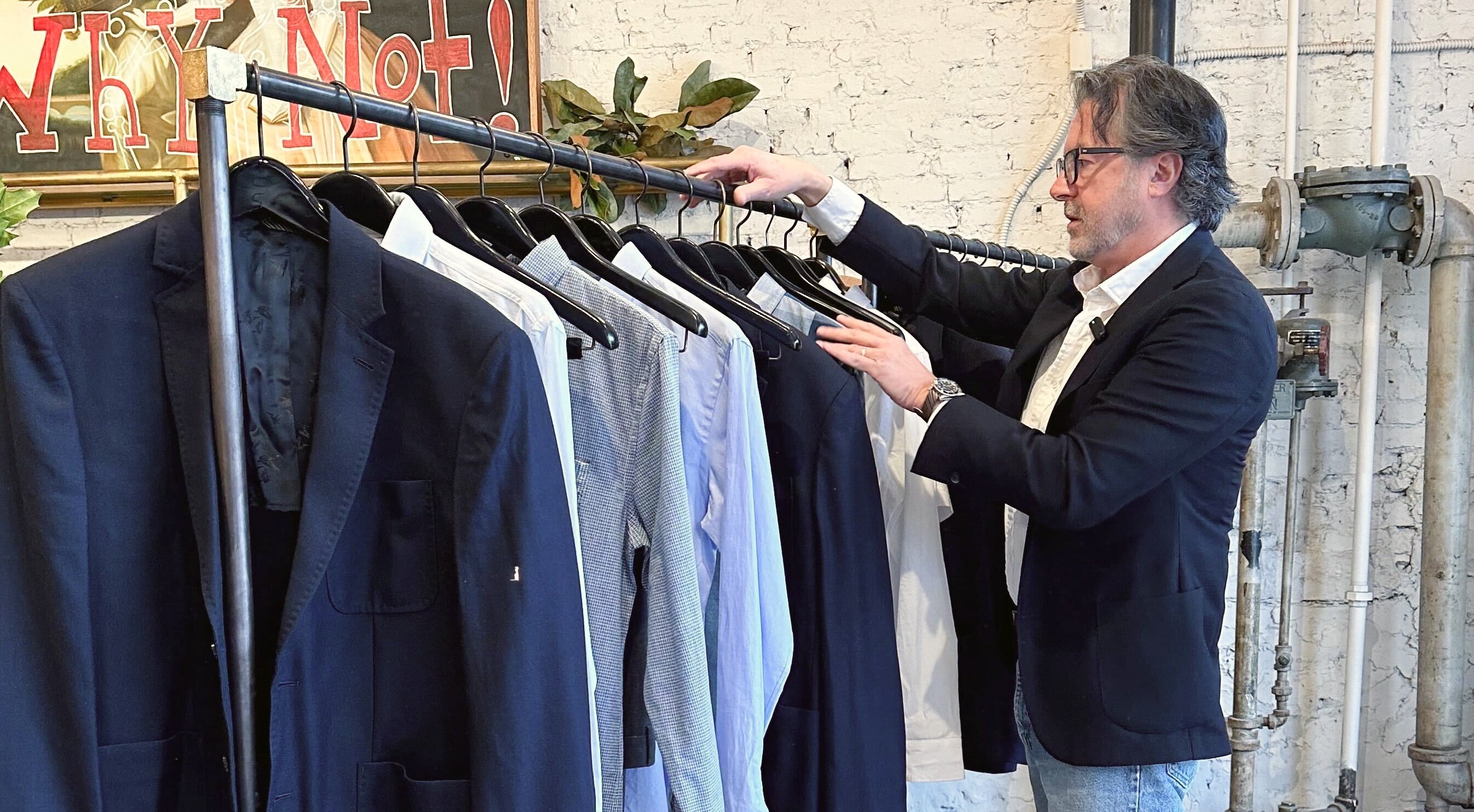 Billy selecting jackets from a clothing rack in-shop with a painting reading "Why Not!" in the background.