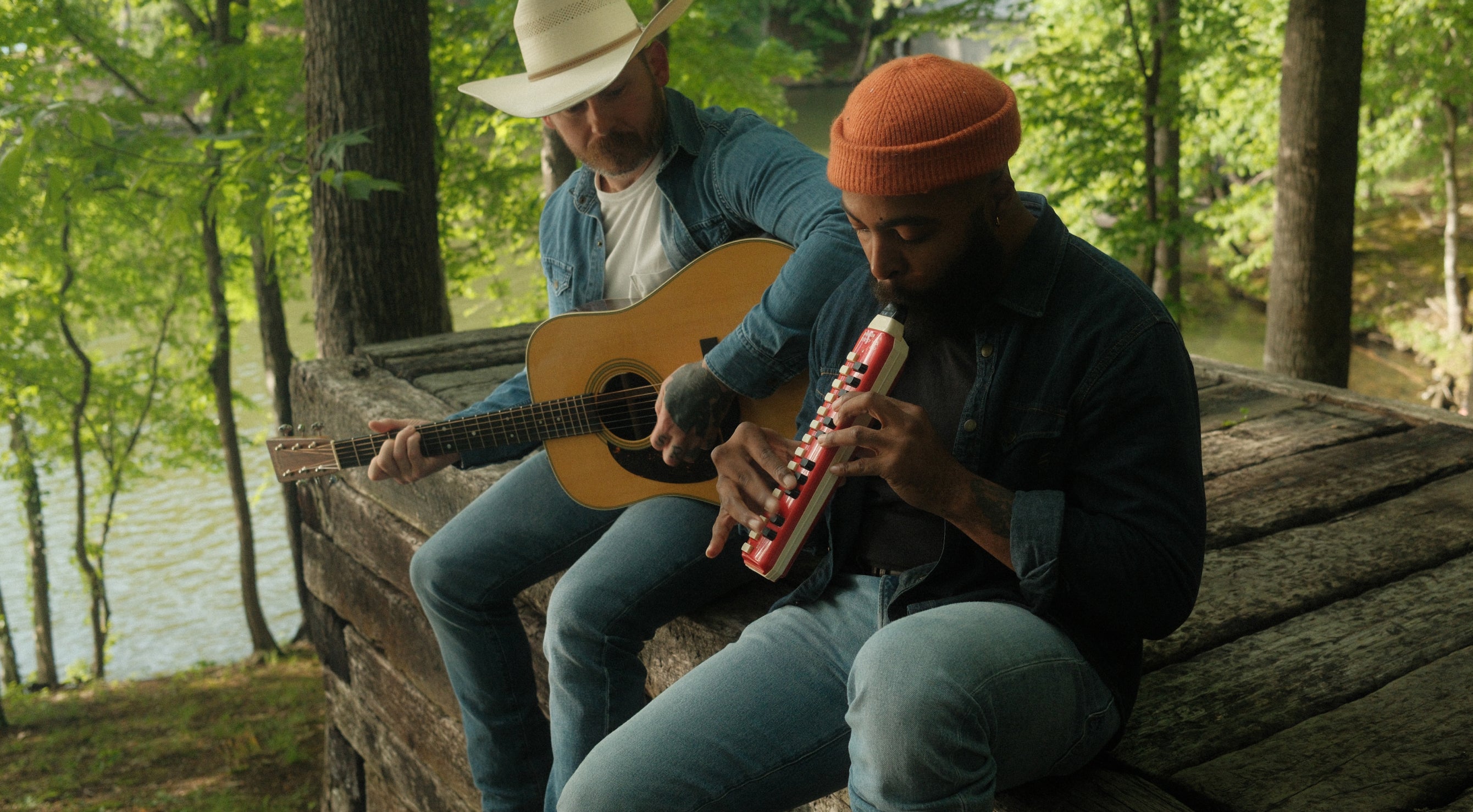 Two people playing guitar and melodica by a lake with trees in the background