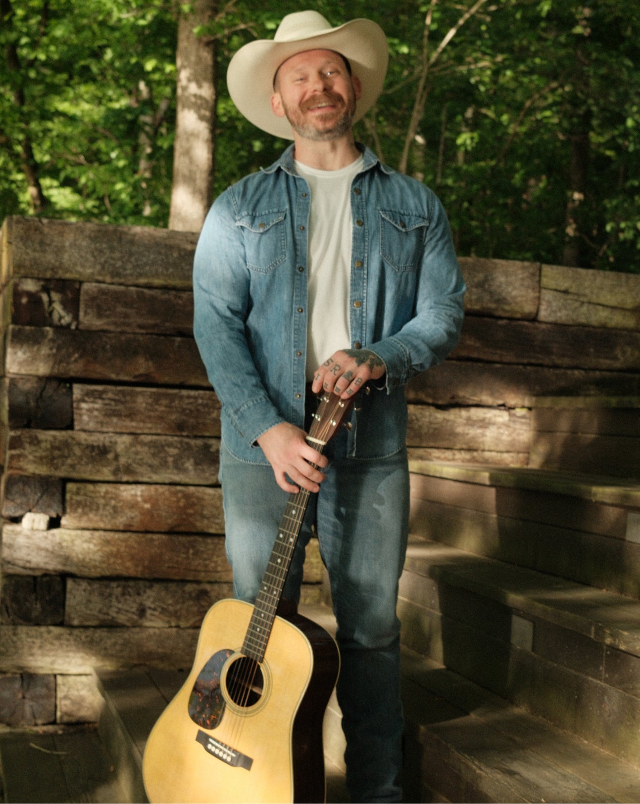 Man in denim jacket and cowboy hat holding a guitar outdoors with wooden steps and greenery.