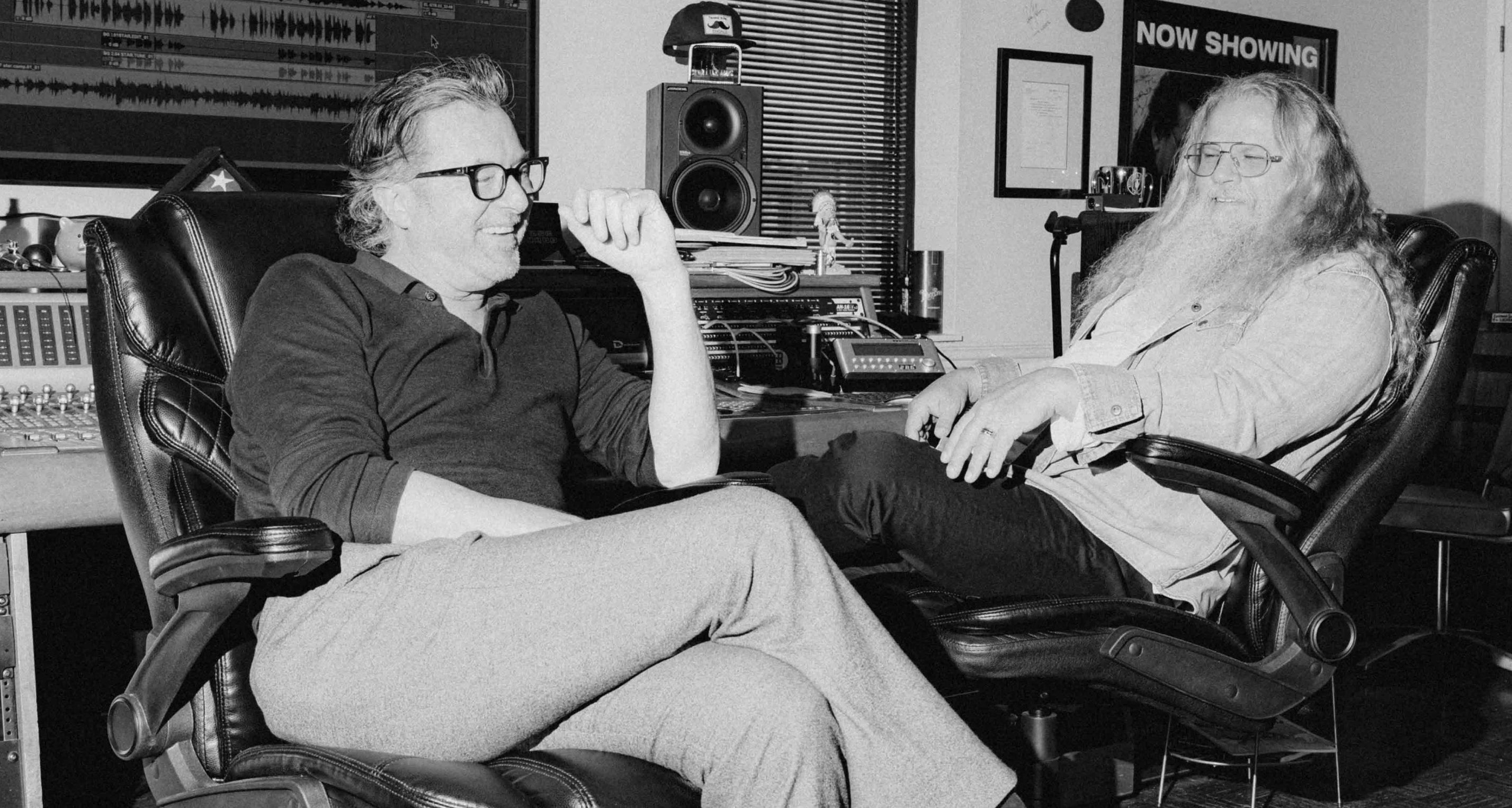 Black and white photograph of two men sitting in leather recording studio chairs, with audio equipment, speakers, and a 'NOW SHOWING' sign visible in the background.