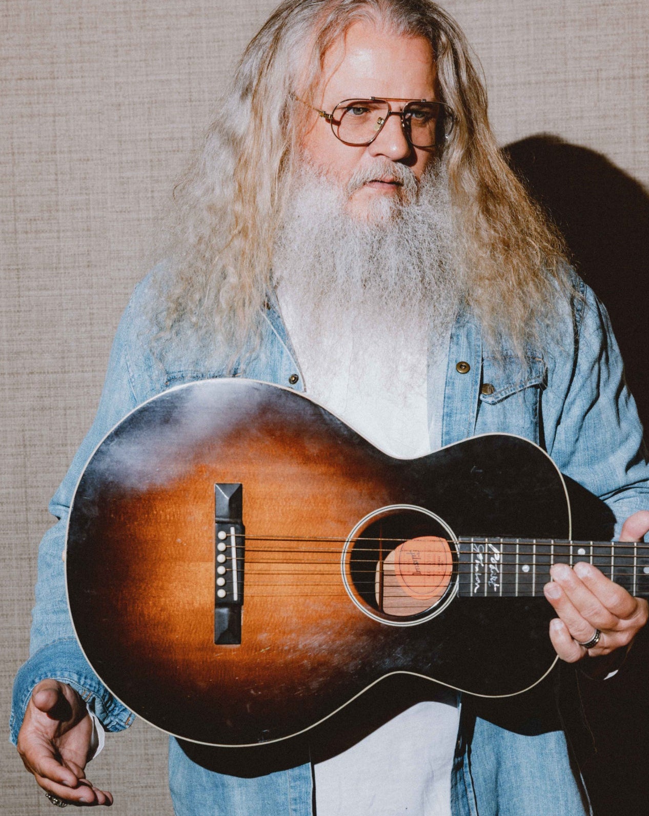 A man with long gray hair and a white beard, wearing glasses and a light blue denim shirt, holding a sunburst acoustic guitar against a textured beige background.