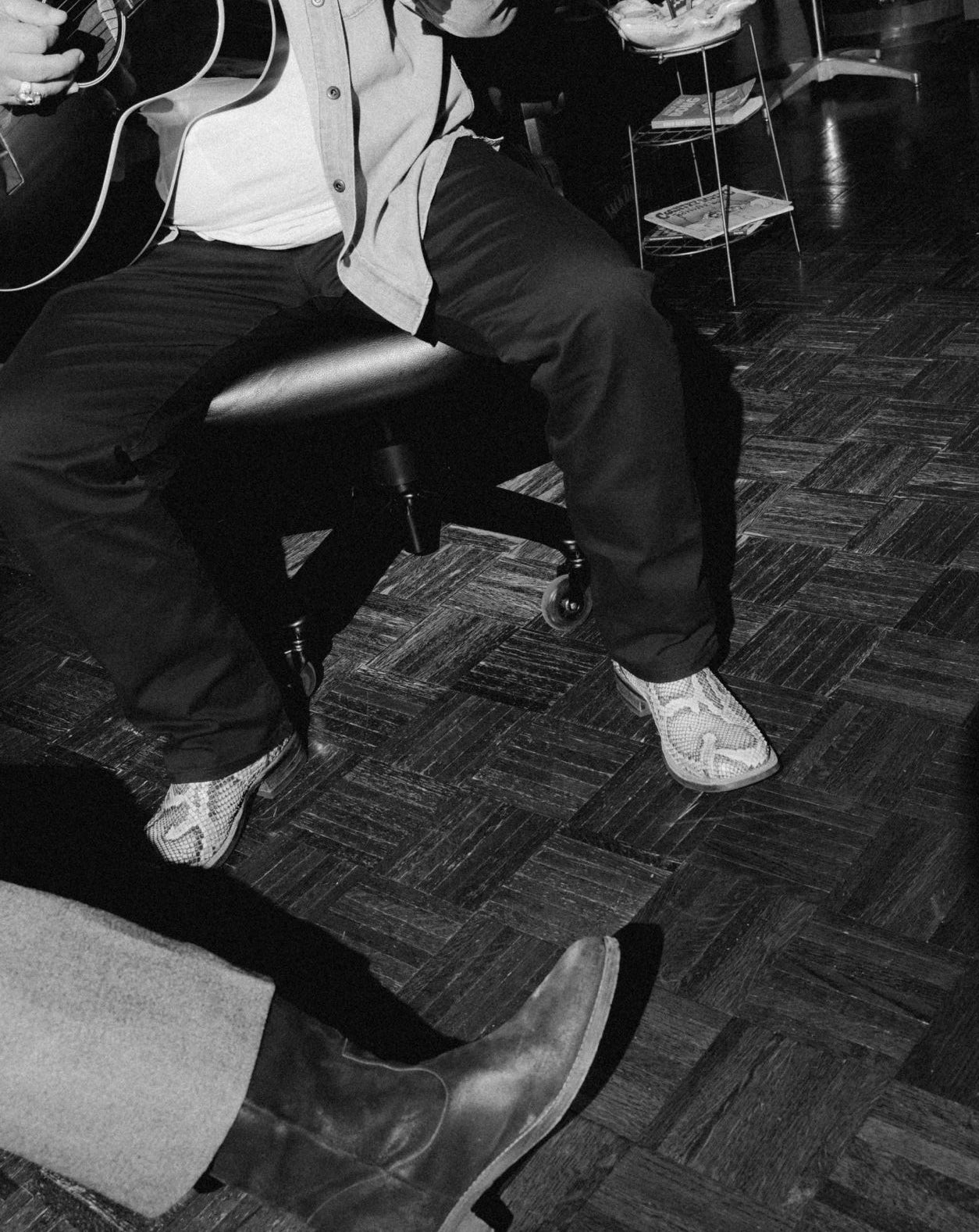 Black and white photograph of a seated person in a room with a wood parquet floor, wearing dark trousers, a light shirt, and light-colored shoes, with another person's boot visible in the foreground.