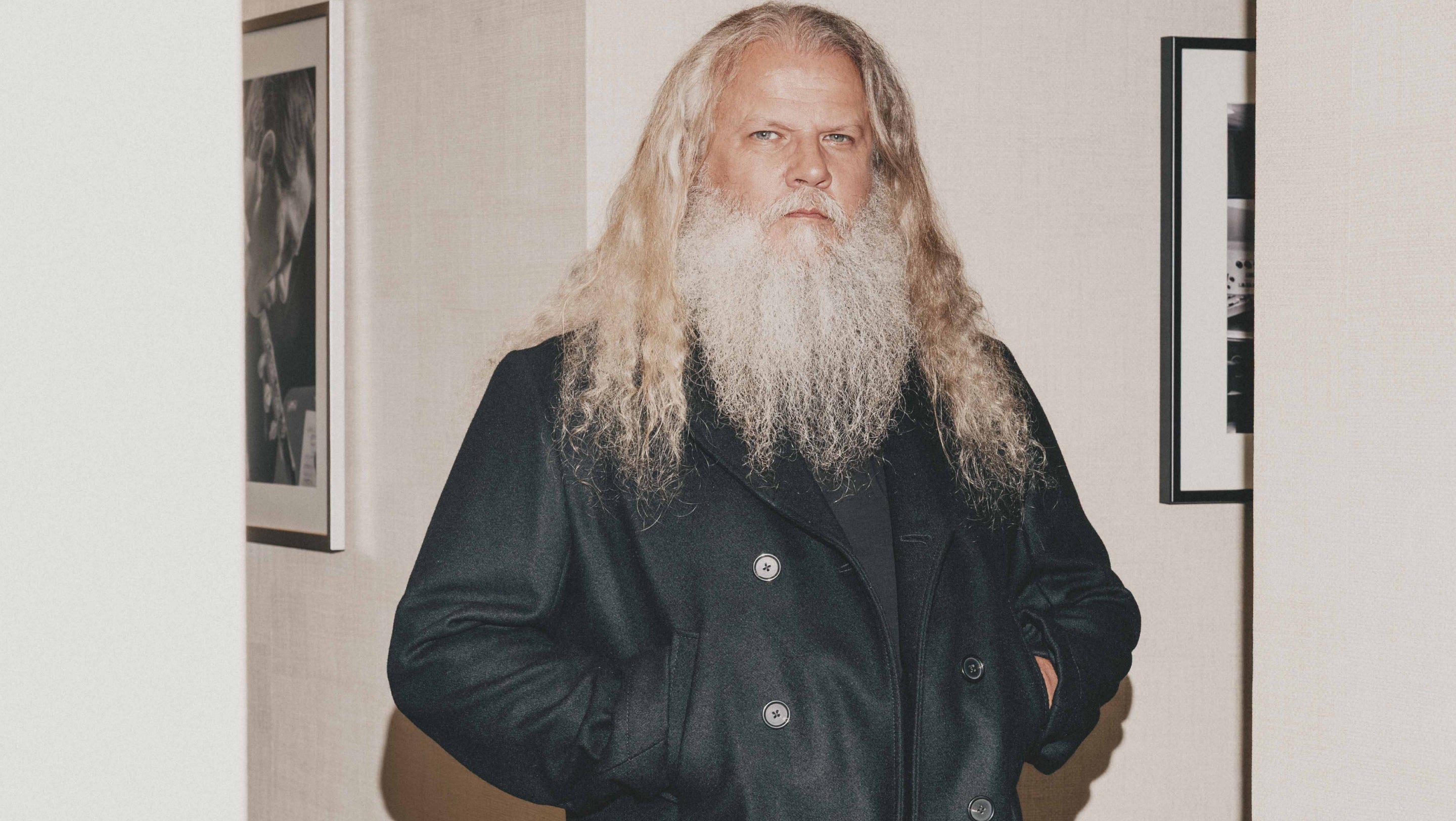 A man with long blonde hair and a full white beard wearing a black double-breasted coat, standing indoors against a light-colored wall with framed pictures.
