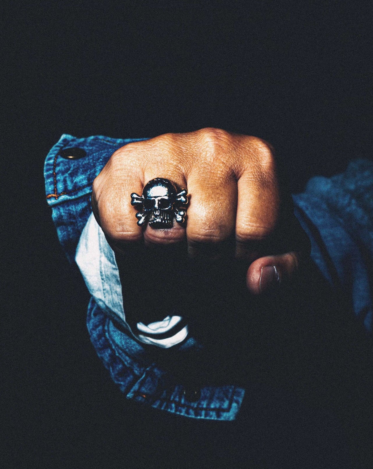 A close-up of a hand wearing a large silver skull ring, with a blue denim jacket sleeve visible against a dark background.