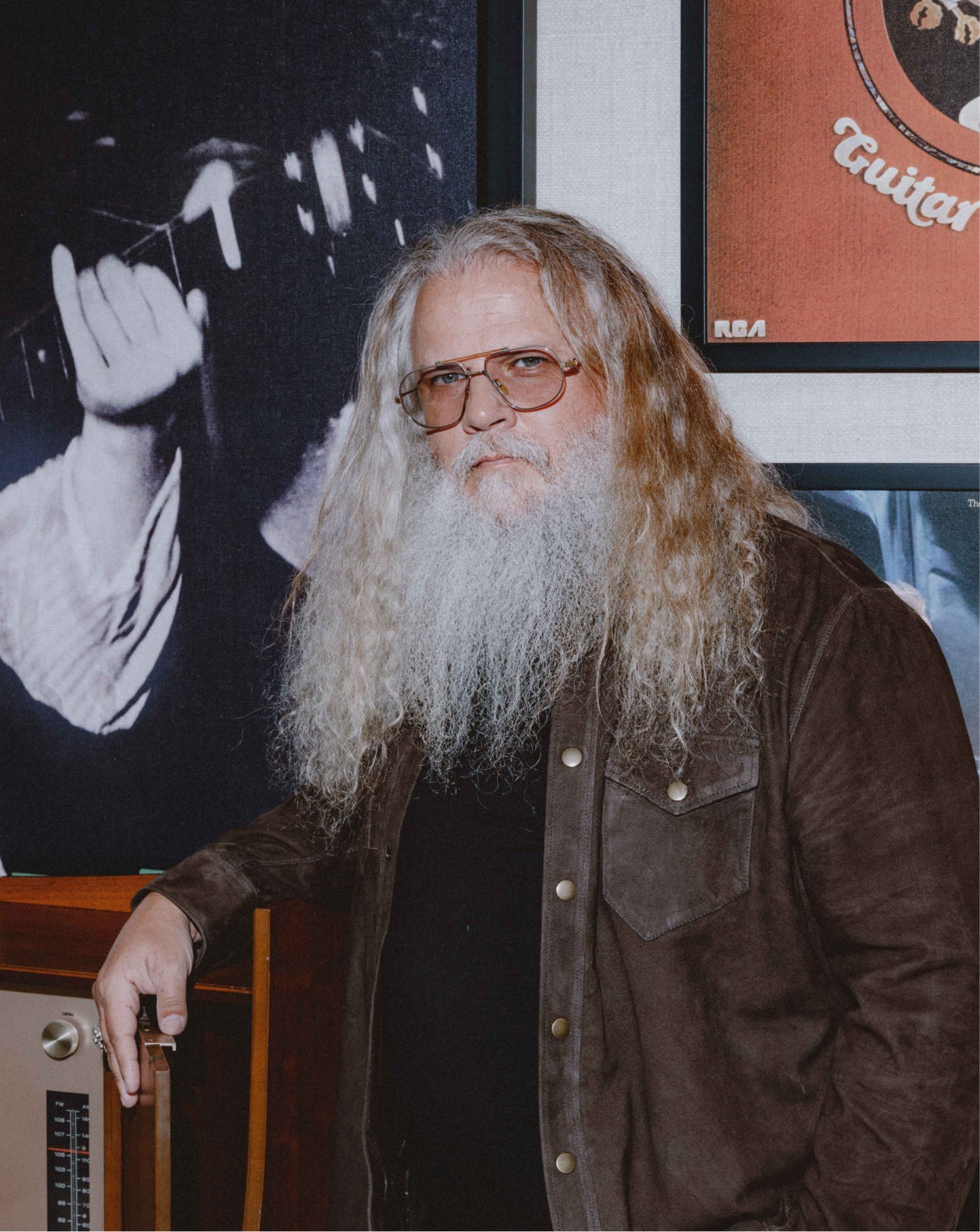A man with a long white beard and glasses, wearing a brown suede jacket over a black shirt, standing in front of framed wall art and a wooden radio console.