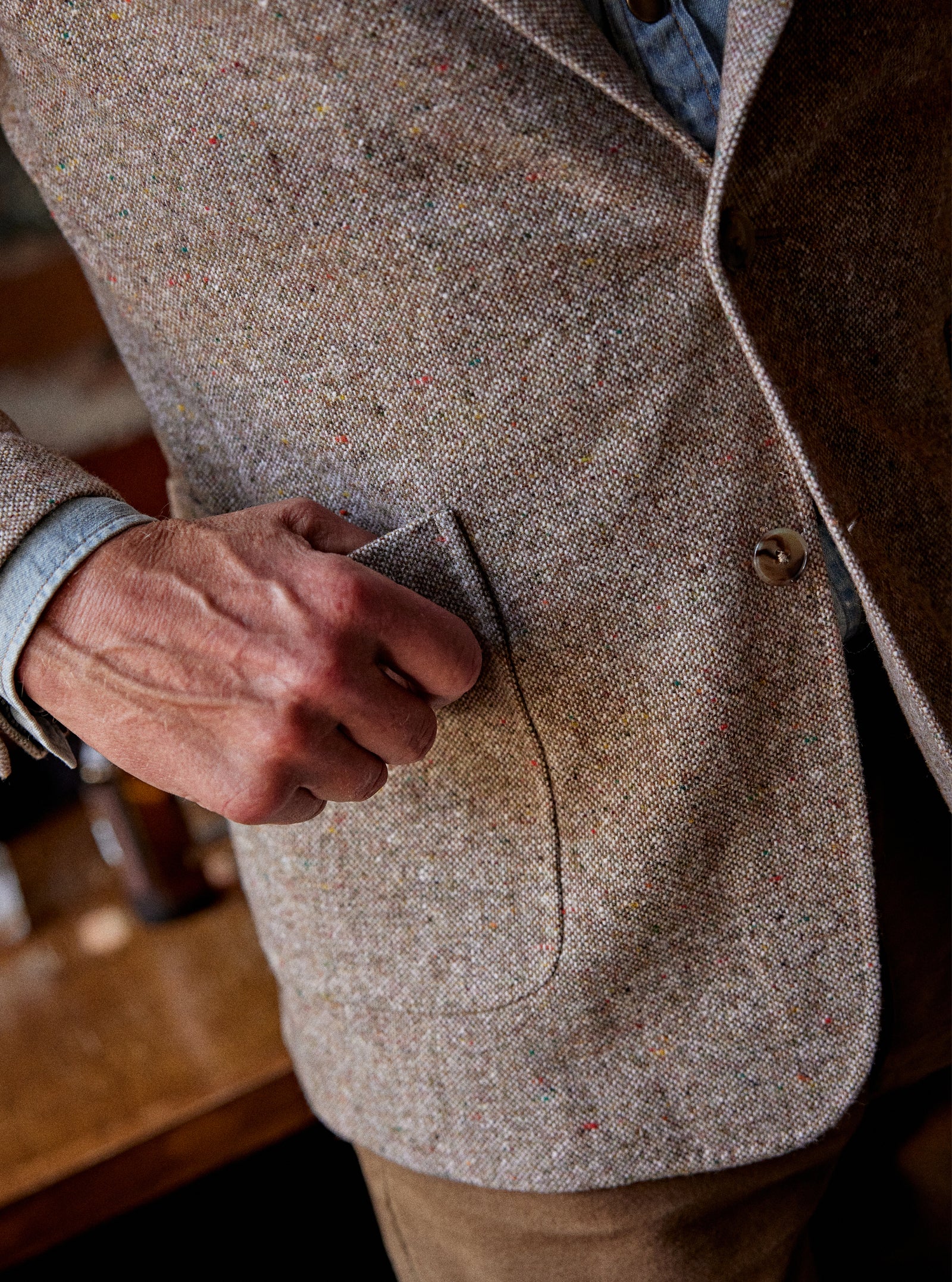Close-up of a person wearing a textured tweed blazer in a warm beige-brown color, with a hand resting on the pocket, set against a softly blurred indoor background.