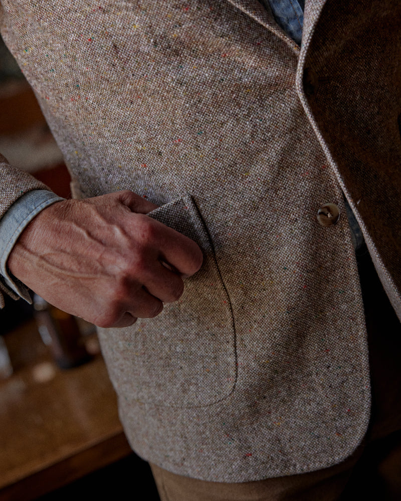 A man with a gray beard stands in a rustic room by a window, wearing a textured beige blazer over a light blue shirt, brown trousers, and dark brown boots.