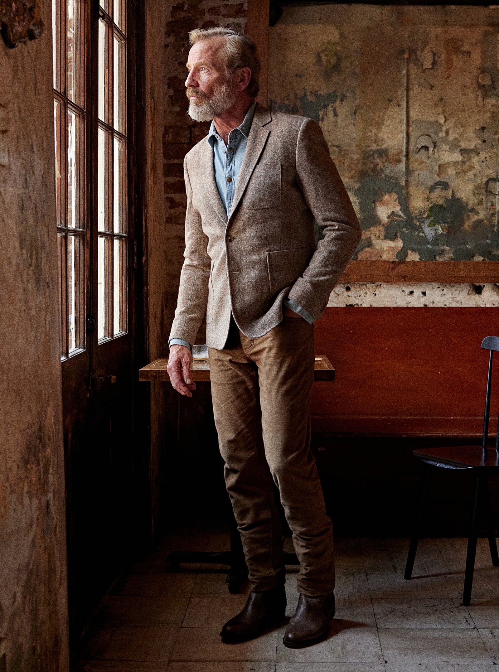 A man with a gray beard stands in a rustic room by a window, wearing a textured beige blazer over a light blue shirt, brown trousers, and dark brown boots.