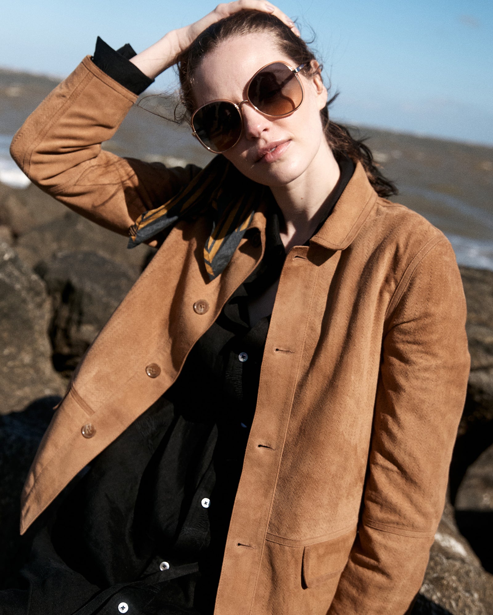 A woman wearing large sunglasses, a tan jacket, and a black shirt stands outside with one hand in her hair. She is in front of large rocks by the water on a sunny day.