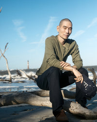 A person with a shaved head, wearing a green long-sleeve shirt and dark pants, sits on driftwood at a beach. They hold a navy cap and look at the camera. The ocean and a distant lighthouse are visible under a blue sky.