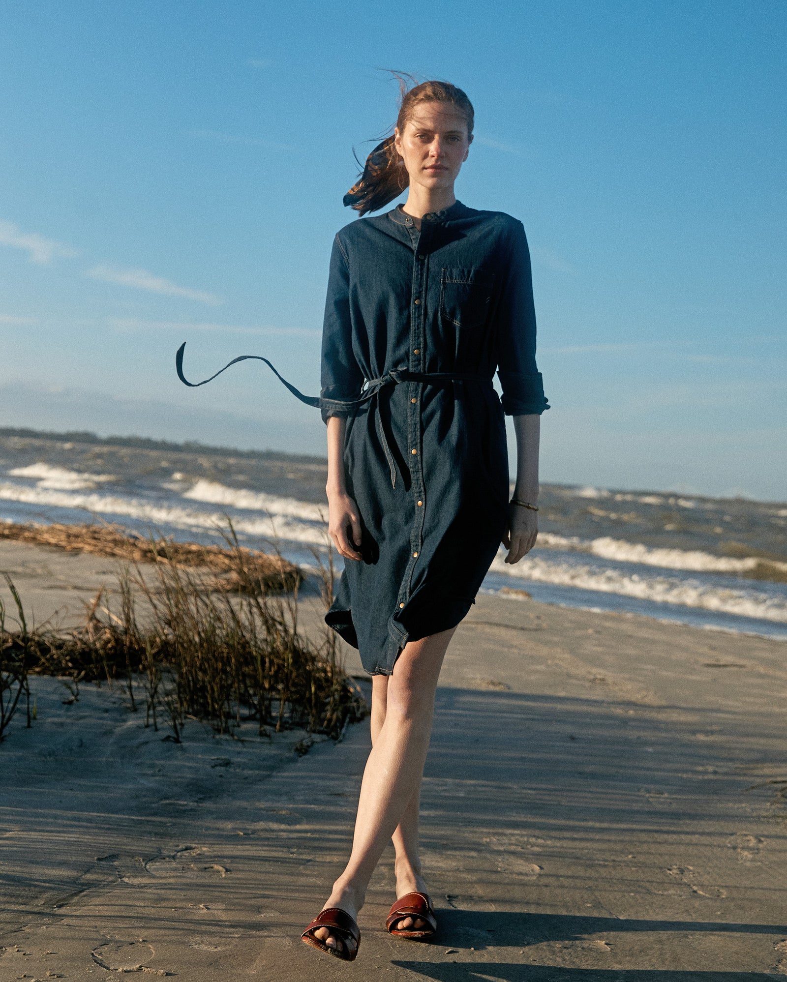 A person in a dark button-up dress stands on a sandy beach with waves in the background and wind blowing their hair and belt. It appears to be a sunny day.