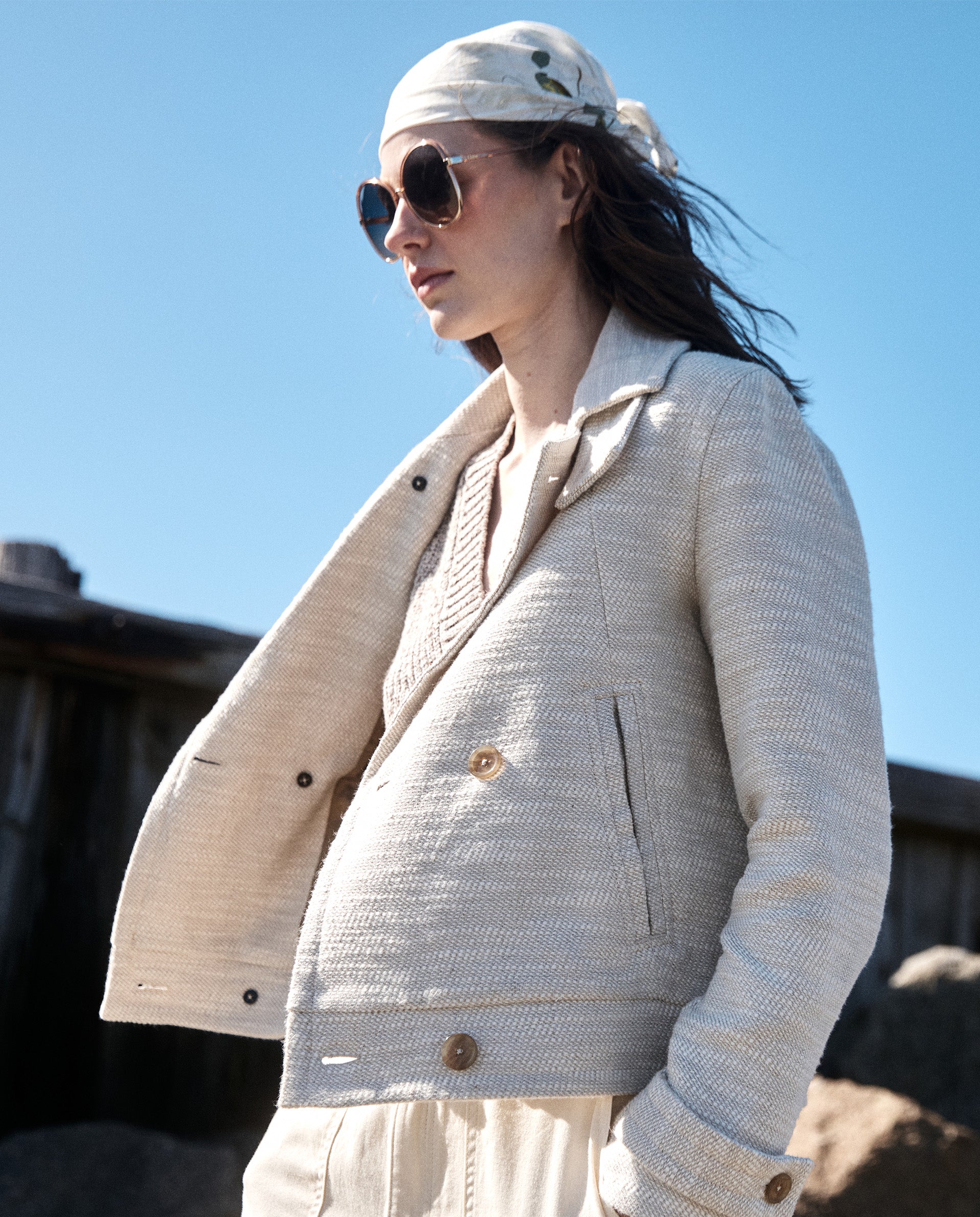 A person wearing sunglasses, a light textured jacket, and a white headscarf stands outdoors in sunlight, with rocks and a wooden fence in the background.