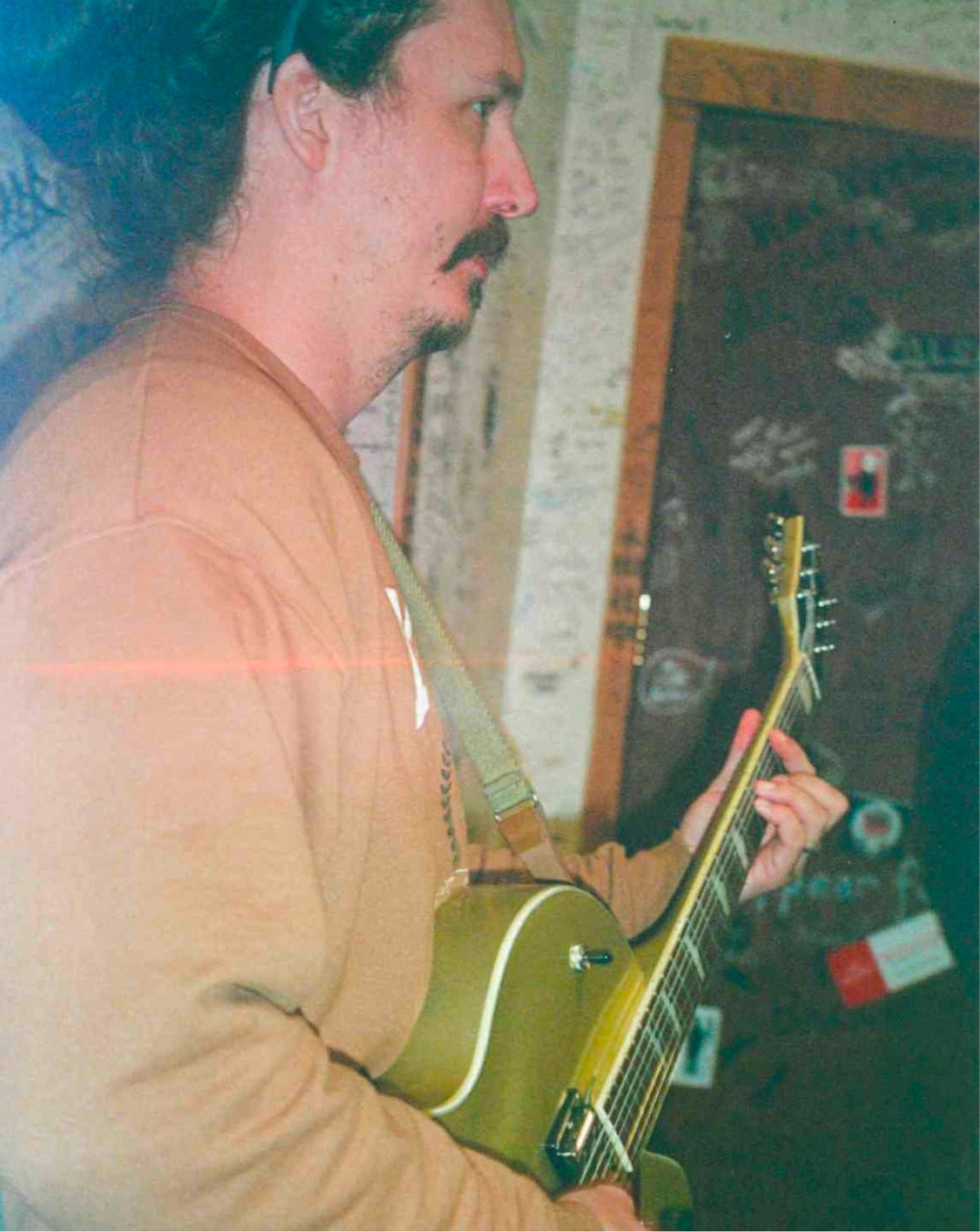 A man in a tan sweatshirt playing an olive green electric guitar in an indoor setting with a graffiti-covered door in the background.
