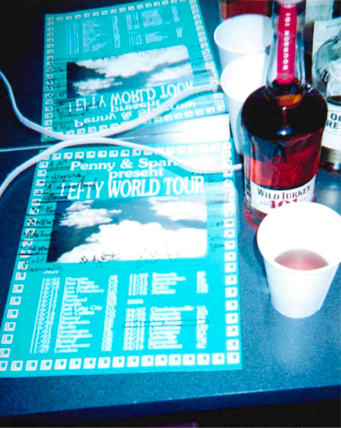A close-up of two printed concert posters for Penny & Sparrow's Lefty World Tour, displayed on a dark table next to a bottle of Wild Turkey 101 and white paper cups.
