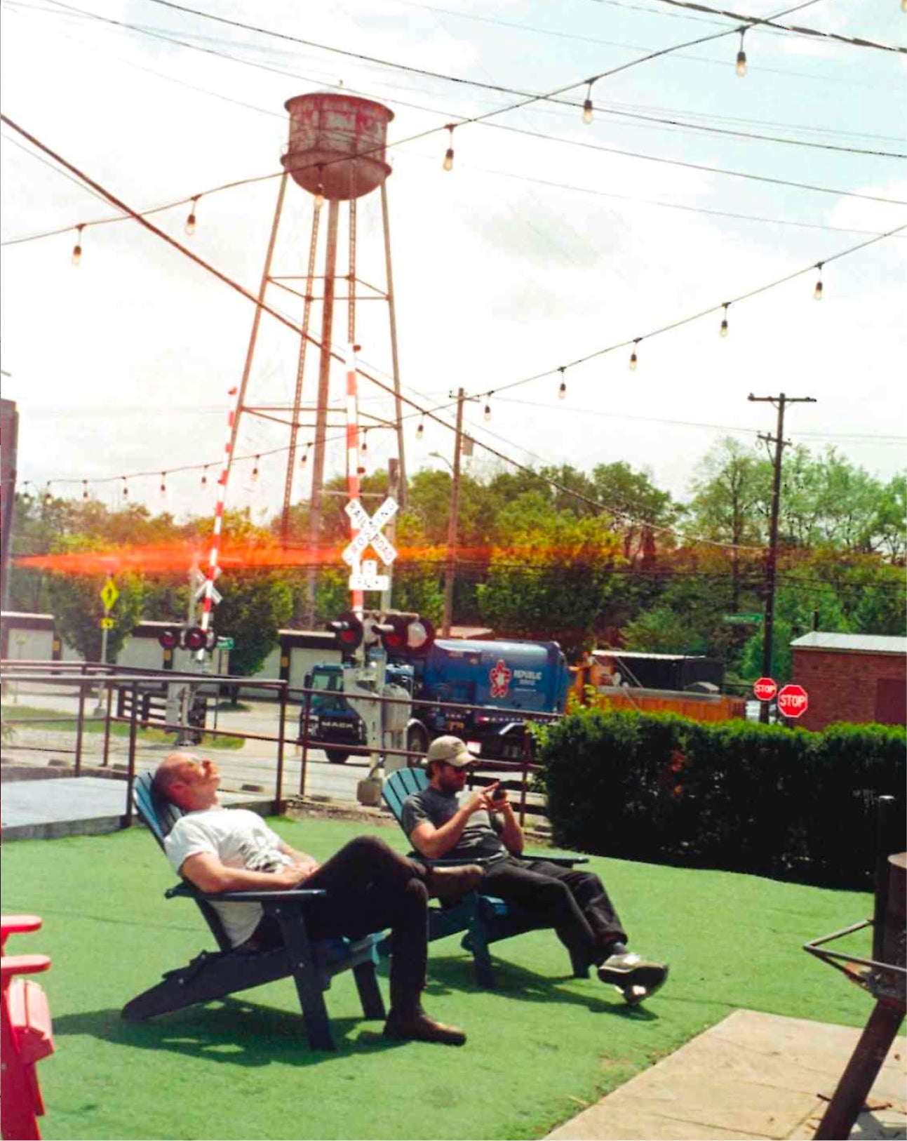 Two people sitting in blue Adirondack chairs on green artificial turf, with a railroad crossing sign and a water tower in the background.