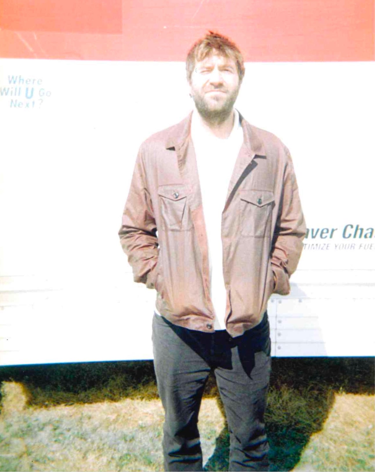A man with a beard wearing a brown jacket, white t-shirt, and dark pants, standing in front of a white truck with a red top banner.