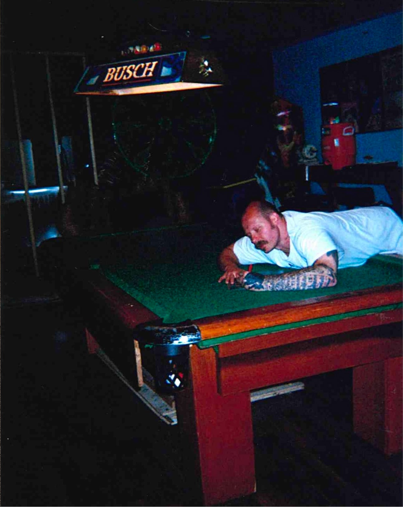 A man in a white t-shirt lies on a green felt pool table in a dimly lit bar or lounge, with a Busch sign visible above and a red cooler in the background.