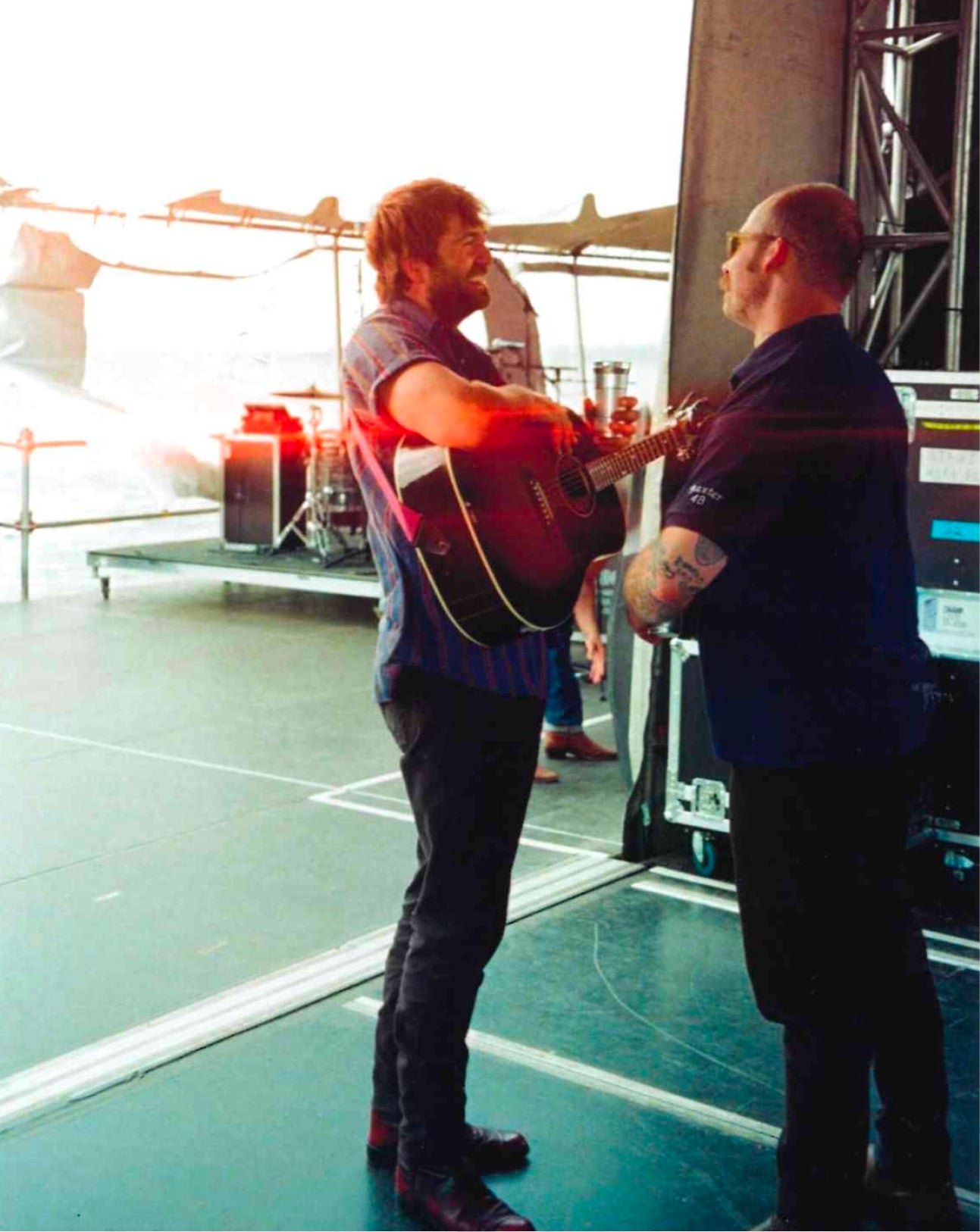 A man in a gray shirt and black pants holds a red acoustic guitar on a stage, facing another man in a dark blue shirt. Bright stage lighting creates a strong flare on the left, and stage equipment is visible in the background.