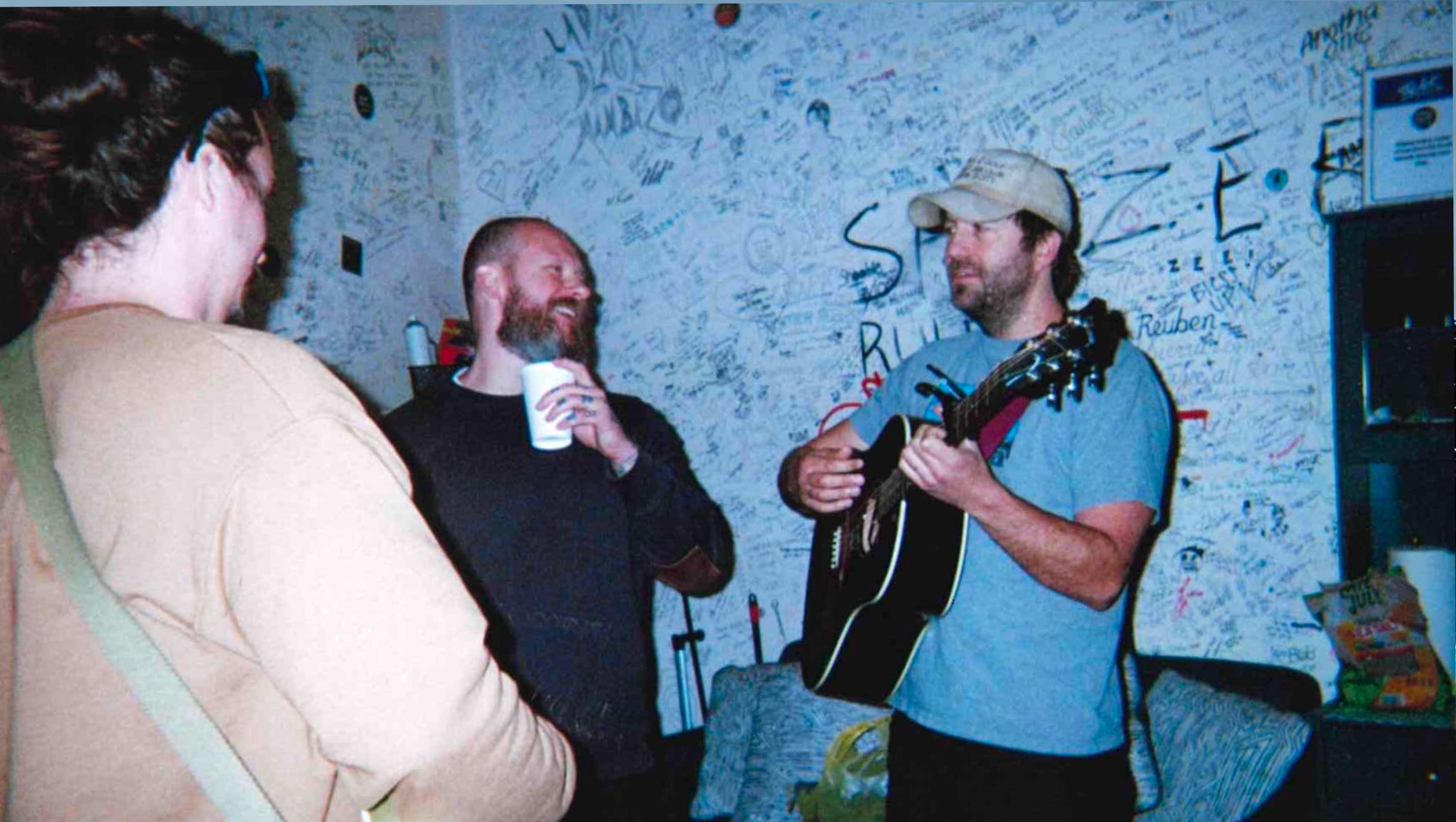 Three people in a casual indoor setting. One man on the right holds a black acoustic guitar and wears a light blue t-shirt and a beige cap.