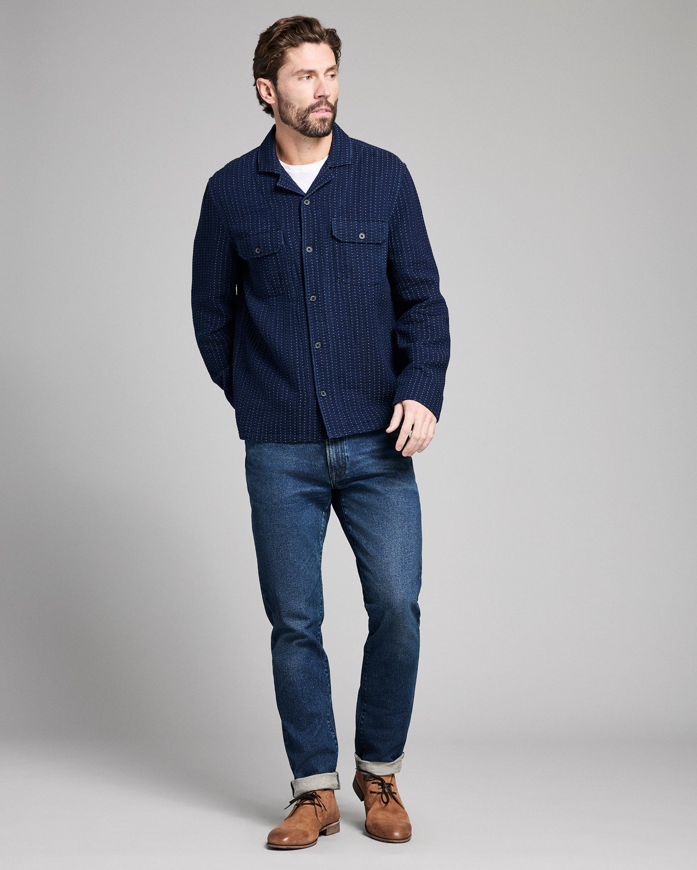 Man wearing a navy blue textured shirt, jeans, and brown shoes on a gray background
