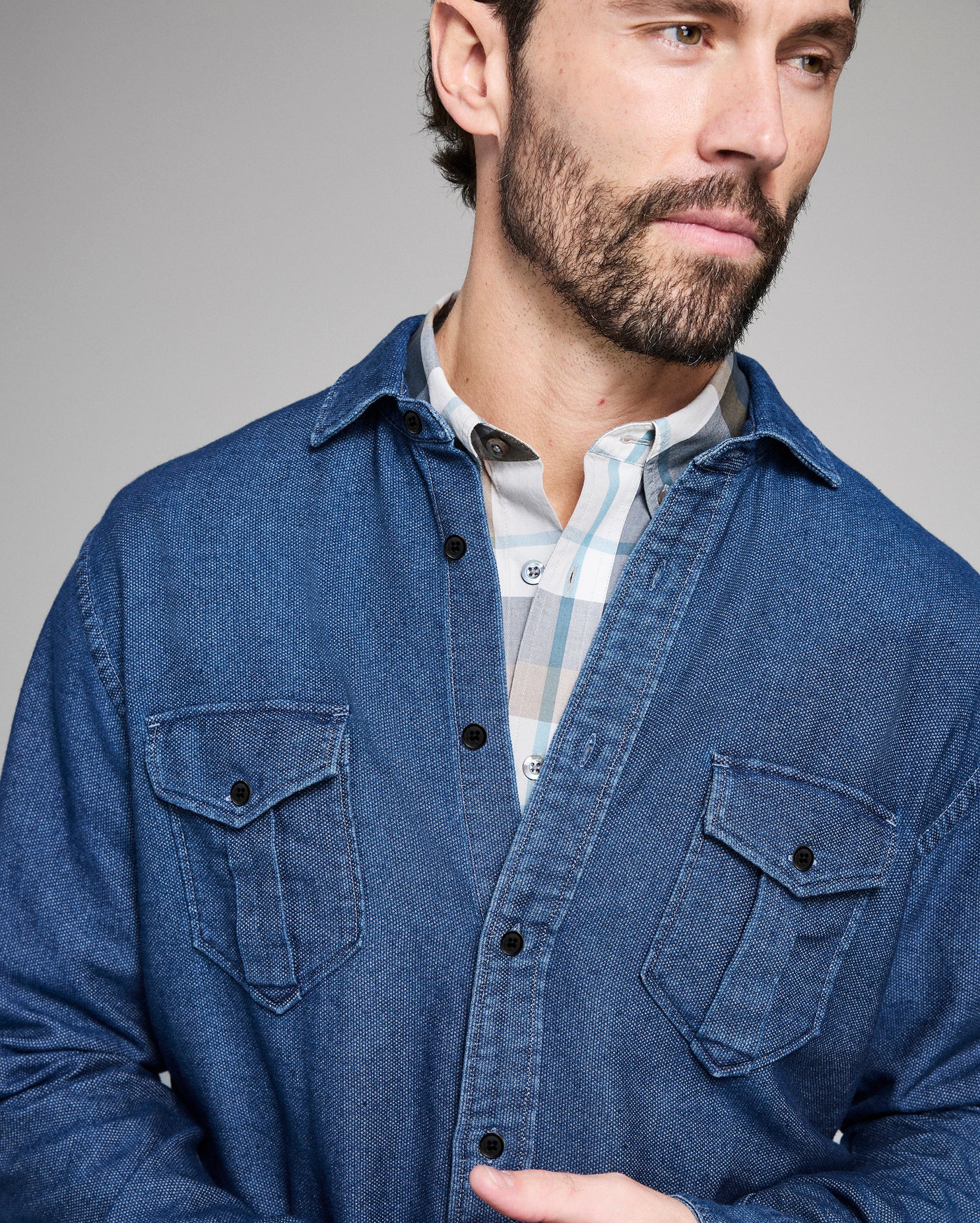 Man wearing a blue denim shirt over a checkered shirt against a gray background