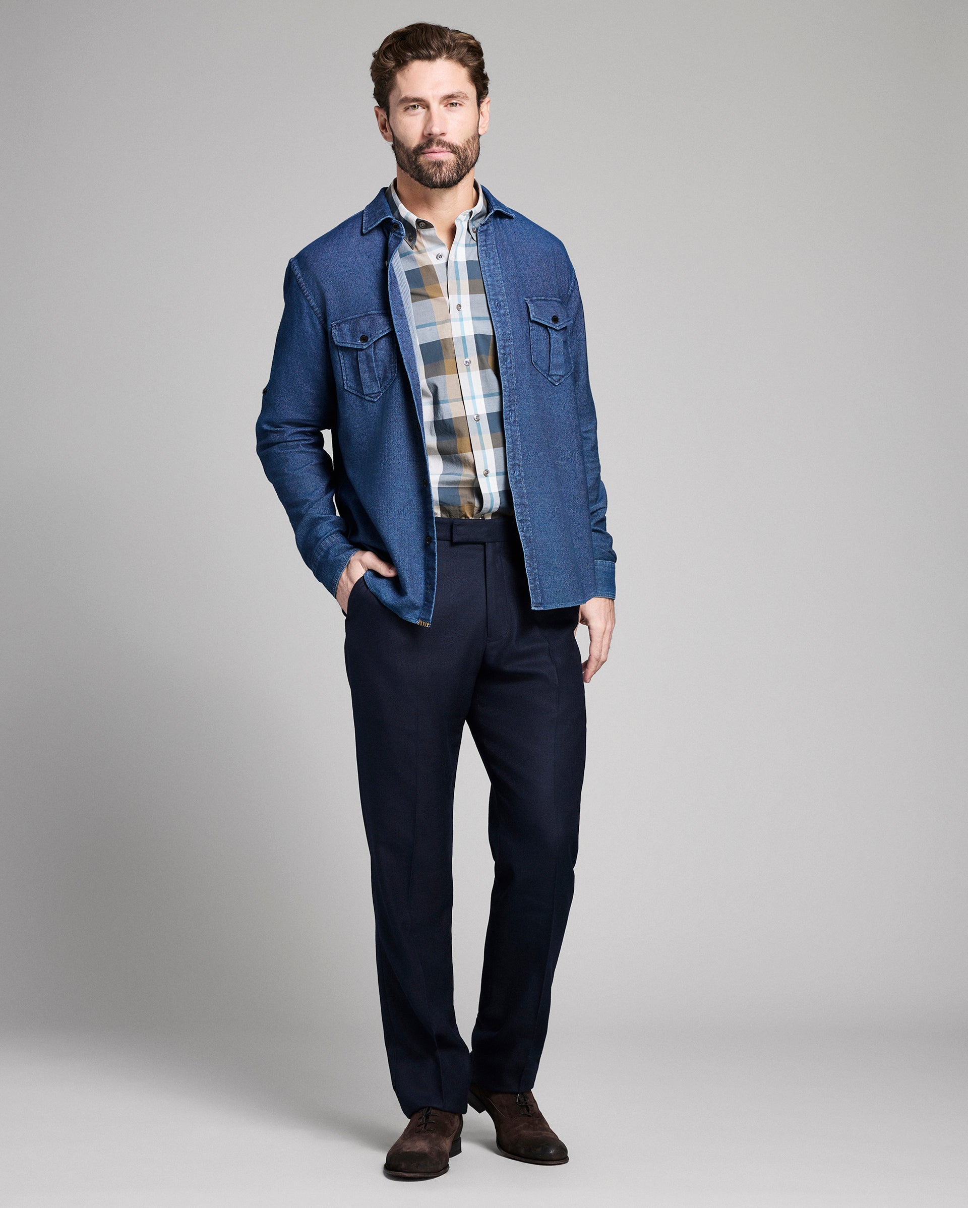 Man wearing a blue denim jacket over a plaid shirt with navy pants on a gray background