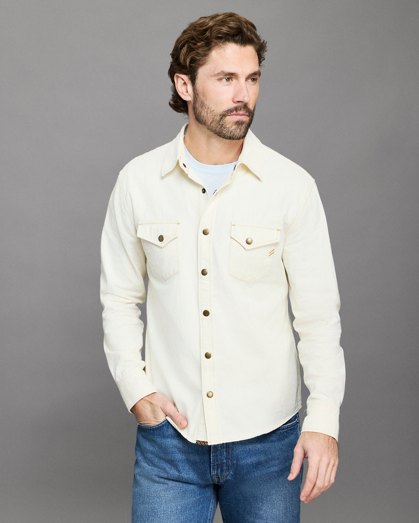 Man wearing a cream-colored denim jacket and blue jeans against a gray background