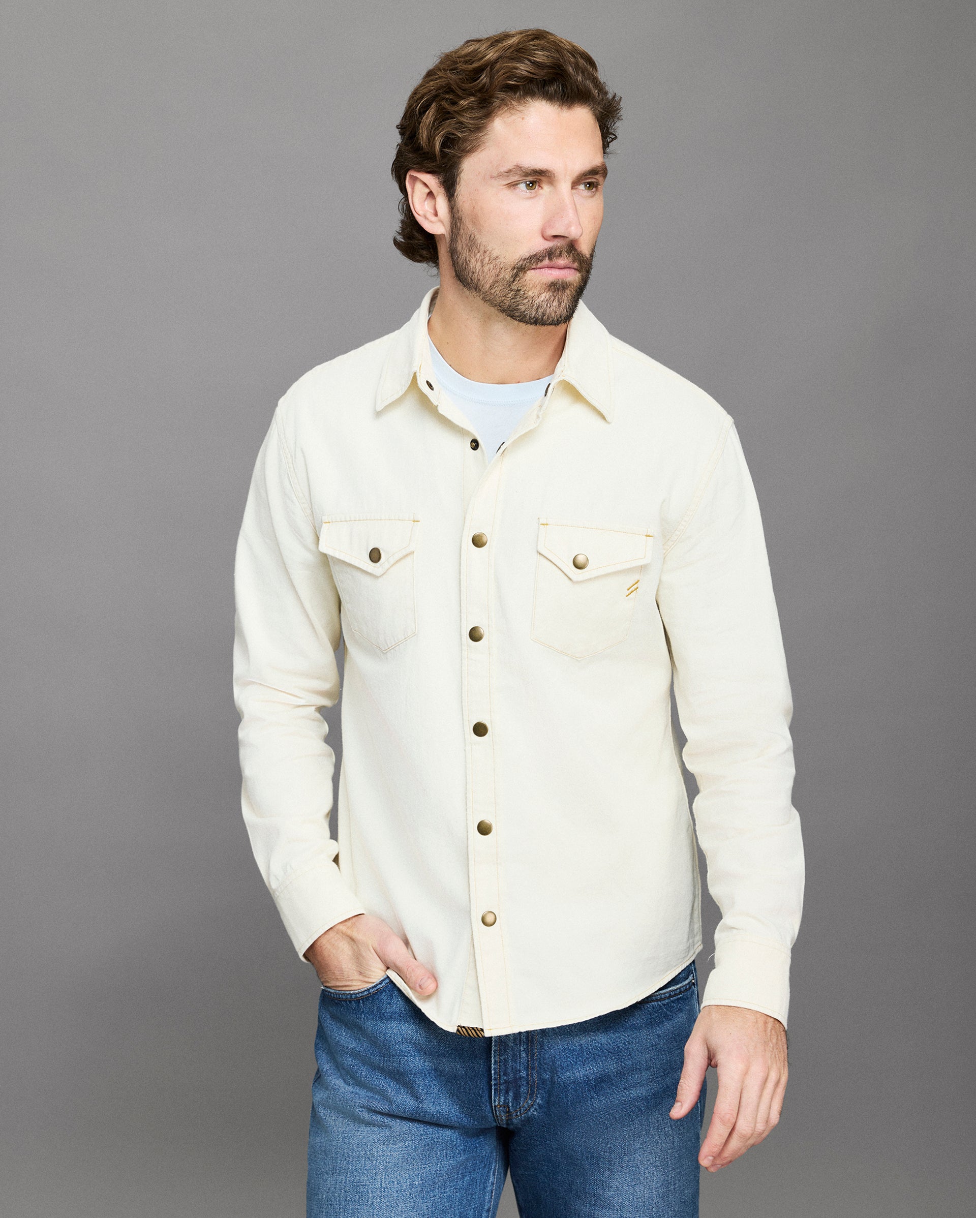 Man wearing a cream-colored denim jacket and blue jeans against a gray background