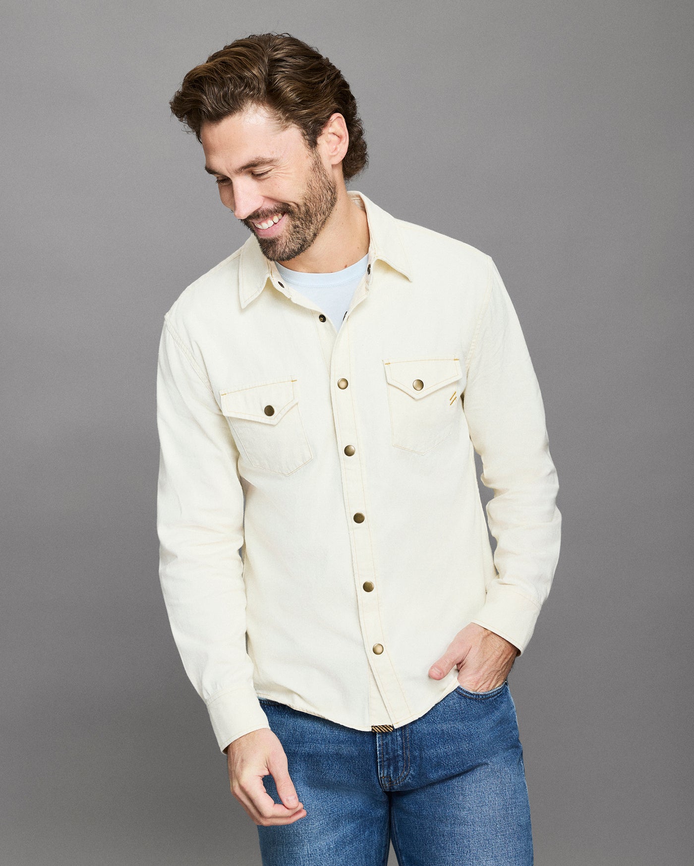 Man wearing a cream denim jacket and blue jeans on a gray background