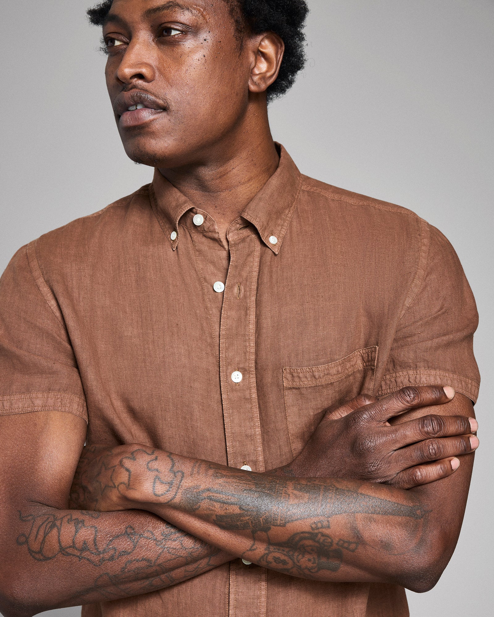 Man wearing a brown shirt with tattoos on his arms against a gray background