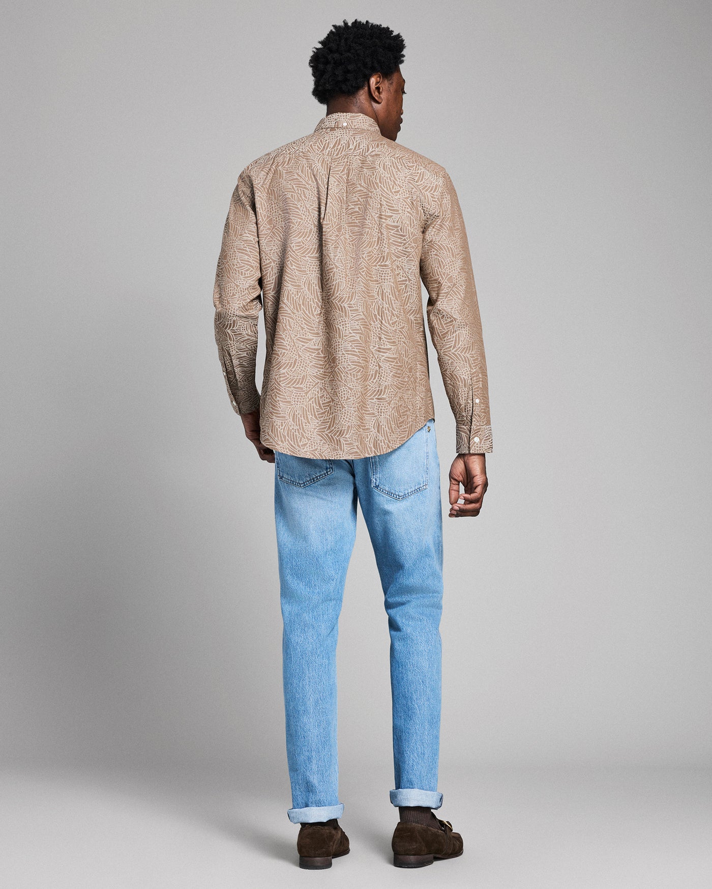 Man wearing a beige patterned shirt and light blue jeans on a gray background