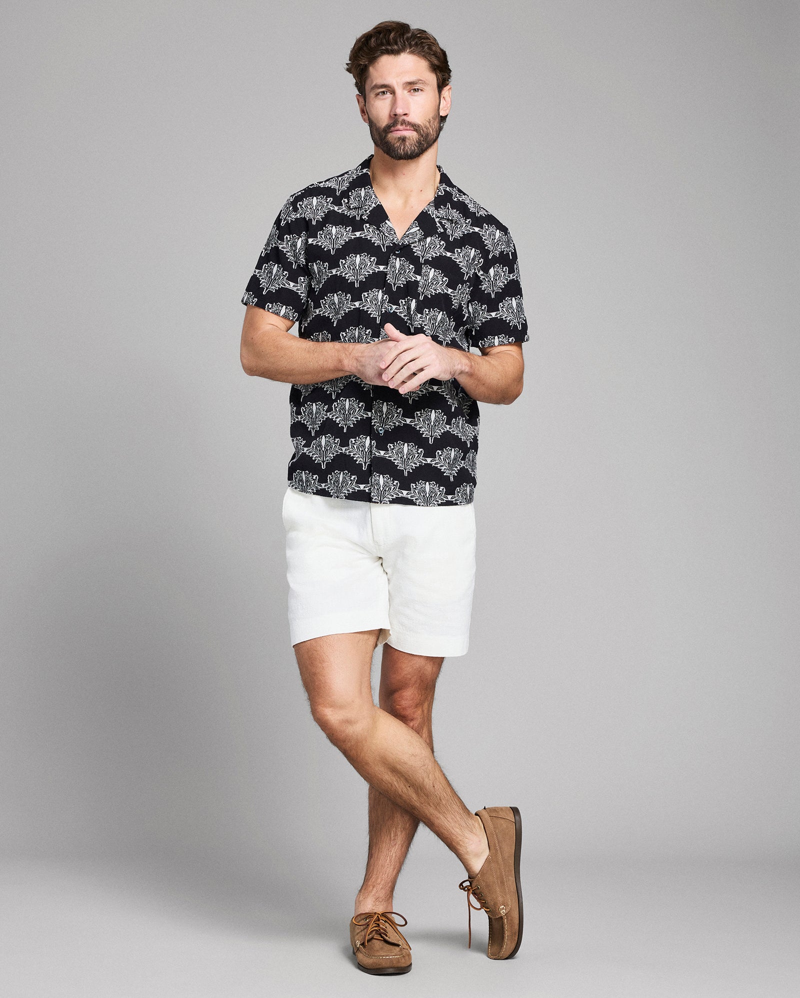 Man wearing a patterned black shirt, white shorts, and brown shoes on a gray background